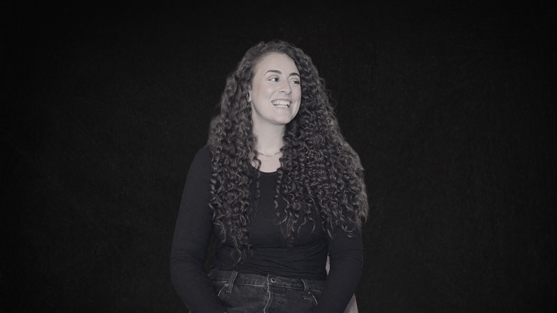 A woman with long, curly hair wearing a black shirt and jeans, smiling while looking to her left against a dark background.
