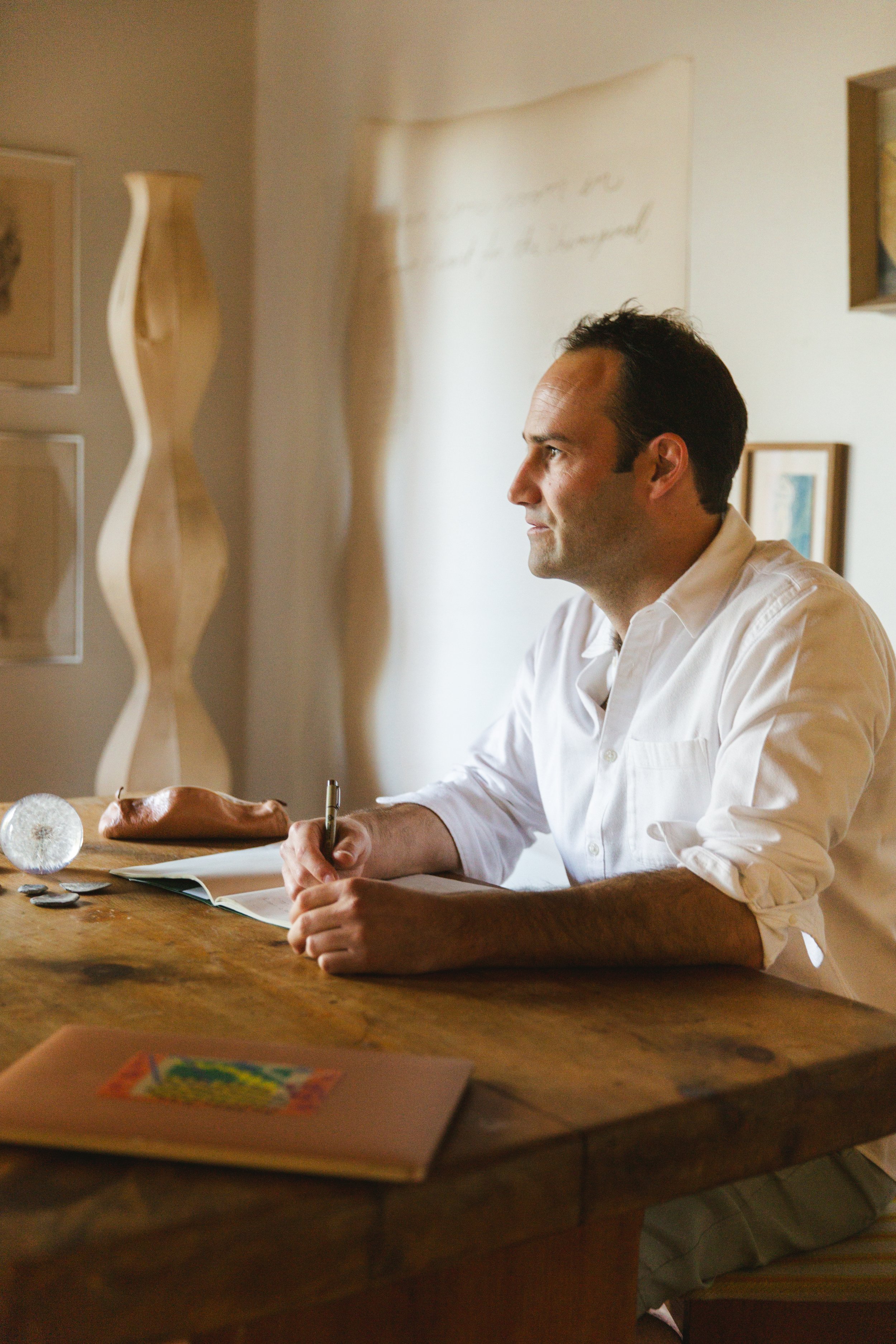 A man with dark hair, wearing a white shirt, sitting at a wooden table with a pen in hand, looking to the right. There is a notebook, some coins, and a leather wallet on the table. The background features a beige wall with framed pictures and a decorative wooden object.