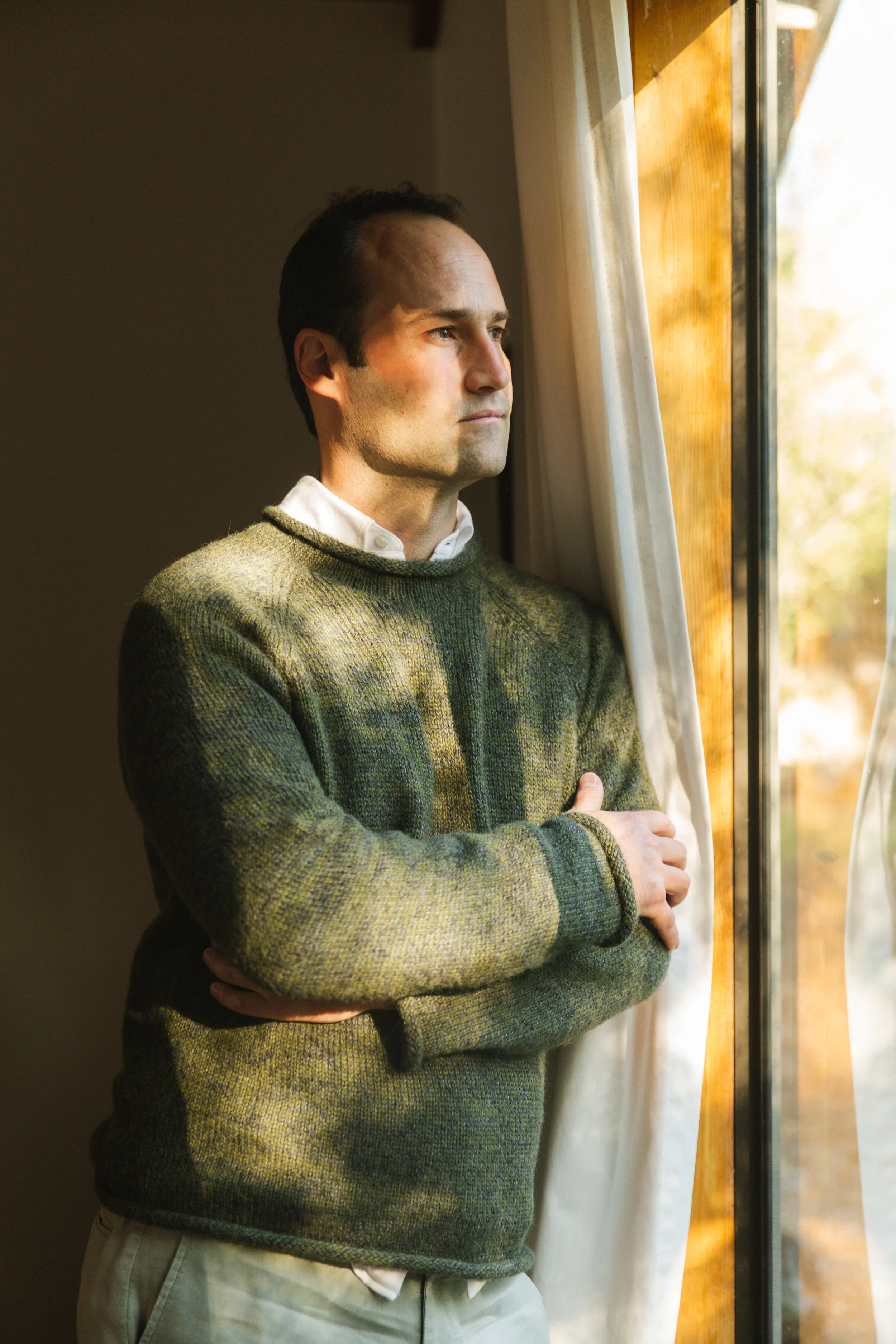 A man standing indoors near a window, looking outside with arms crossed, wearing a greenish-gray sweater and light-colored pants.
