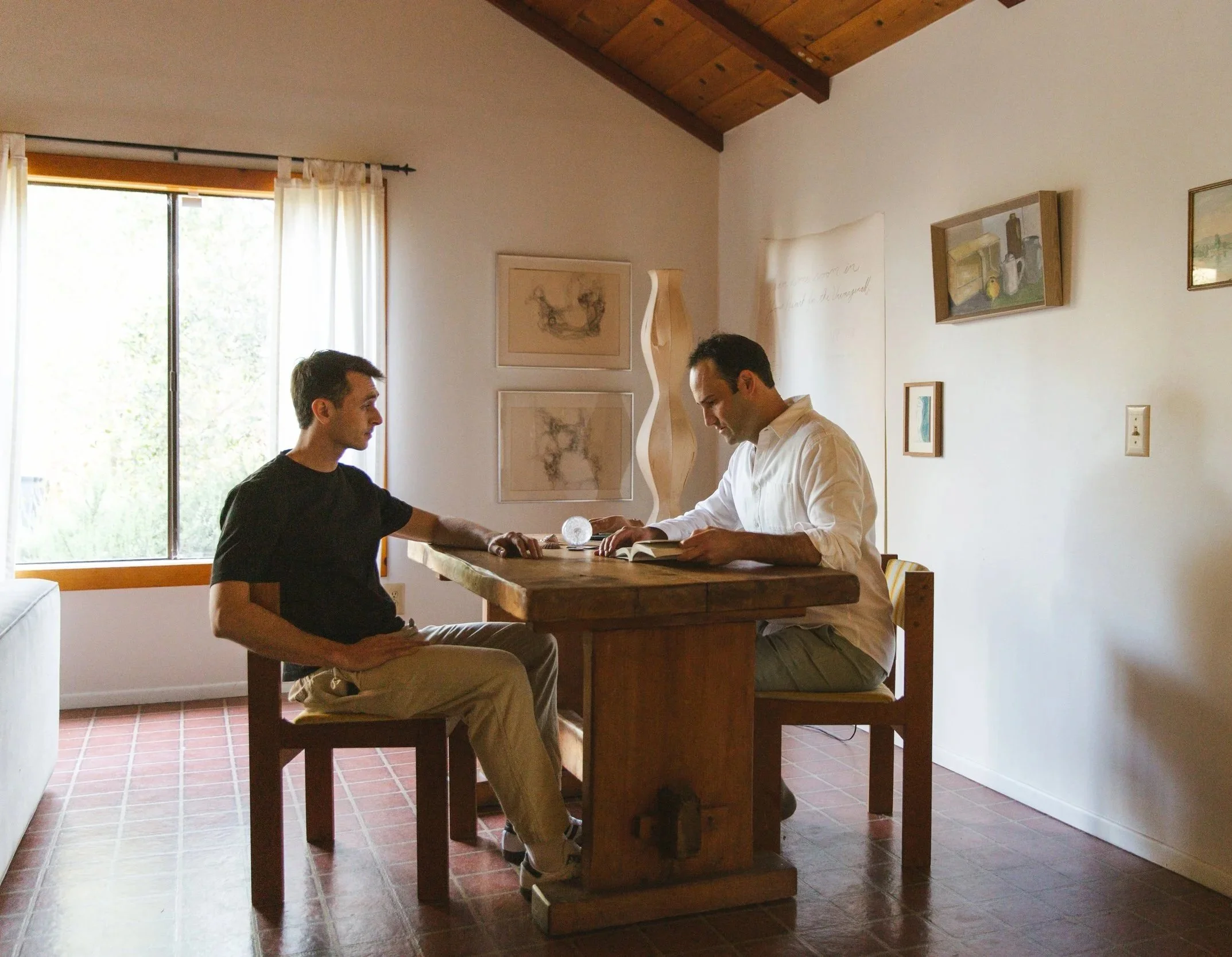 Two men sitting at a wooden dining table in a well-lit room, engaging in conversation with a glowing orb on the table. The room features white walls with framed artwork, a large window with white curtains, and a wooden ceiling.