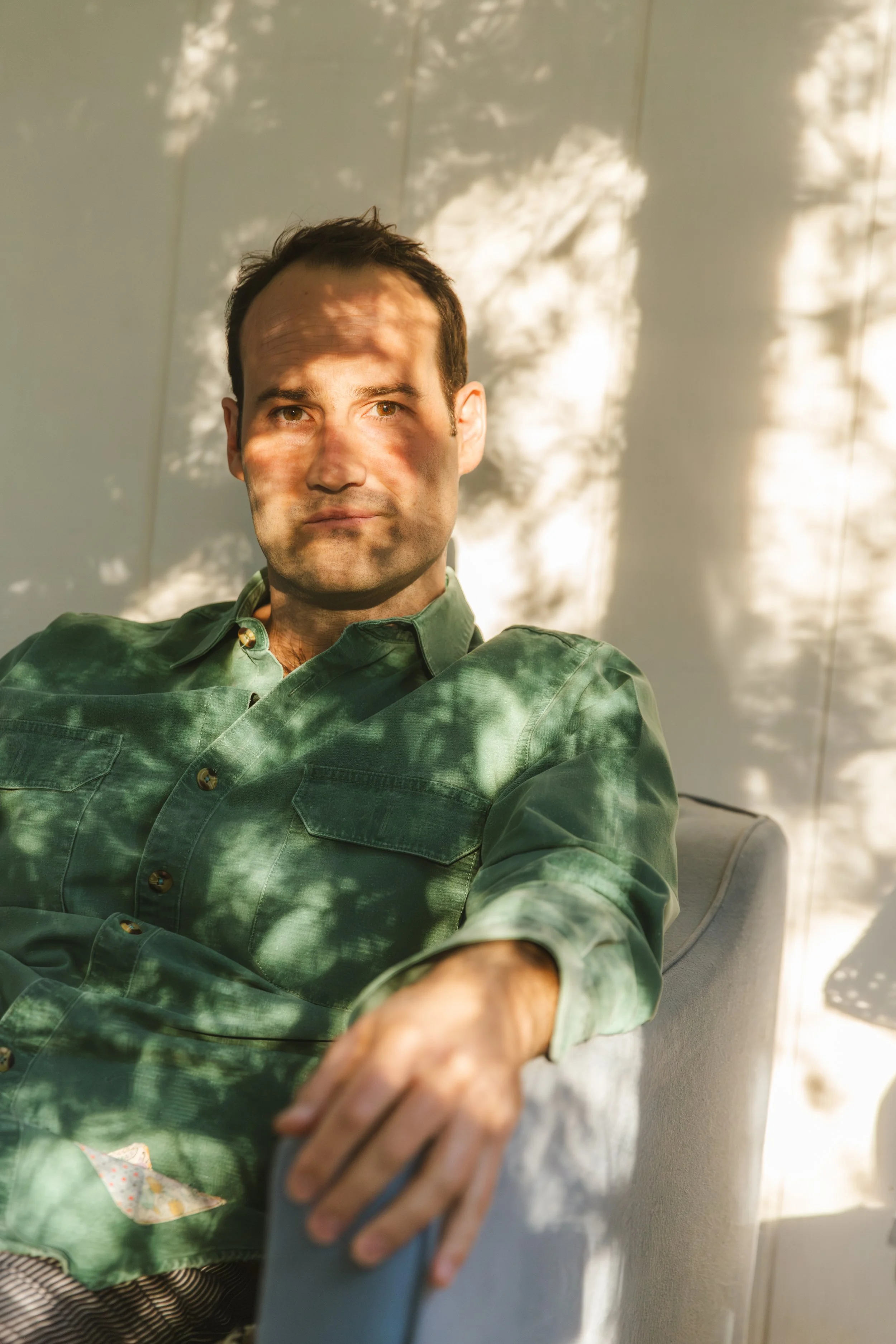 A man with short dark hair and light skin sitting in a chair, wearing a green plaid shirt, with sunlight and shadow patterns on his face and background.