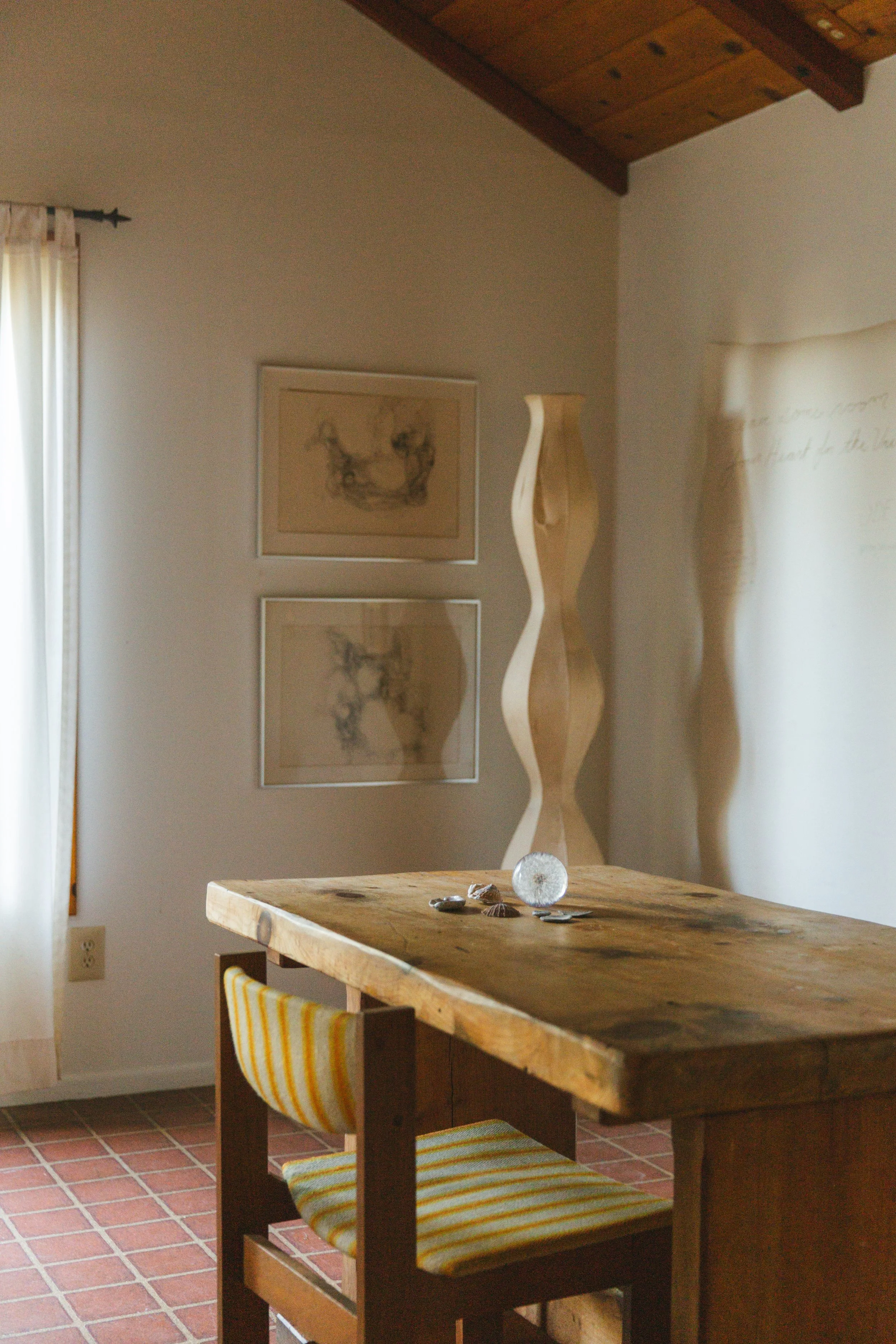 A corner of a room with a wooden ceiling and a brick-tiled floor. There is a wooden table with a striped yellow and white chair. On the table, there are decorative stones and a crystal. The wall has two framed sketches and a tall wavy wooden sculpture. A curtain is partially visible on the left side.