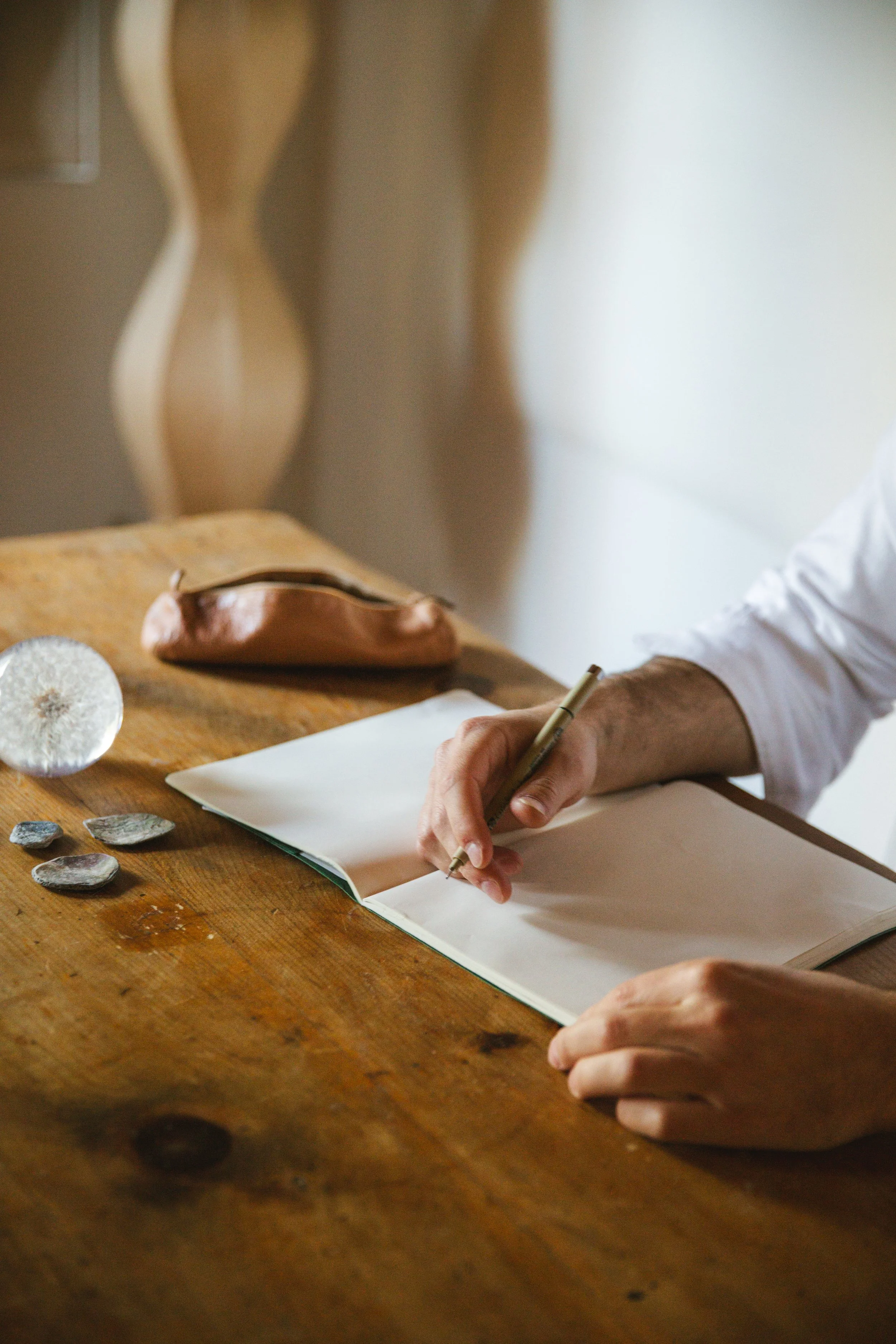 Person writing in a sketchbook at a wooden table with decorative stones and a ceramic object, with blurred furniture in the background.