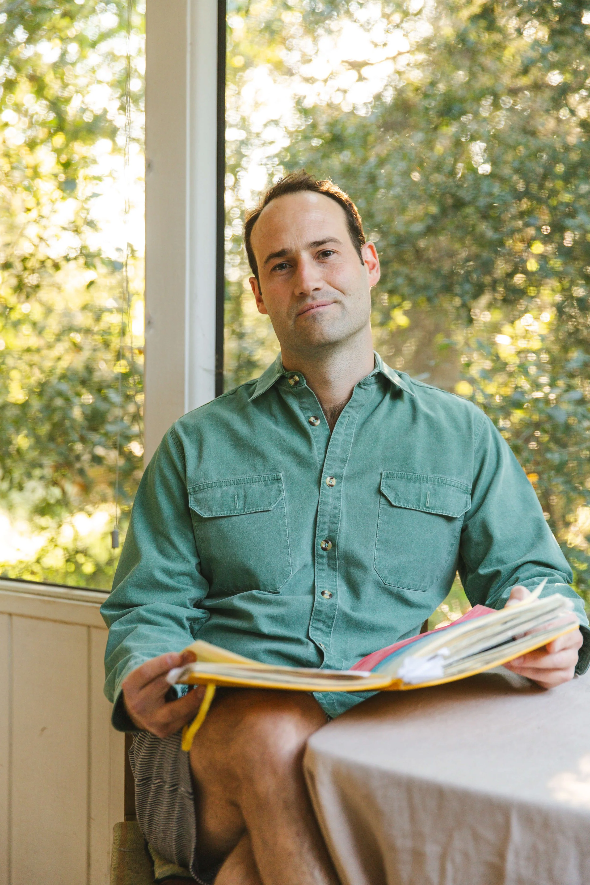 A young man sitting at a table with a book, holding a pen, near a window with trees outside in the background.