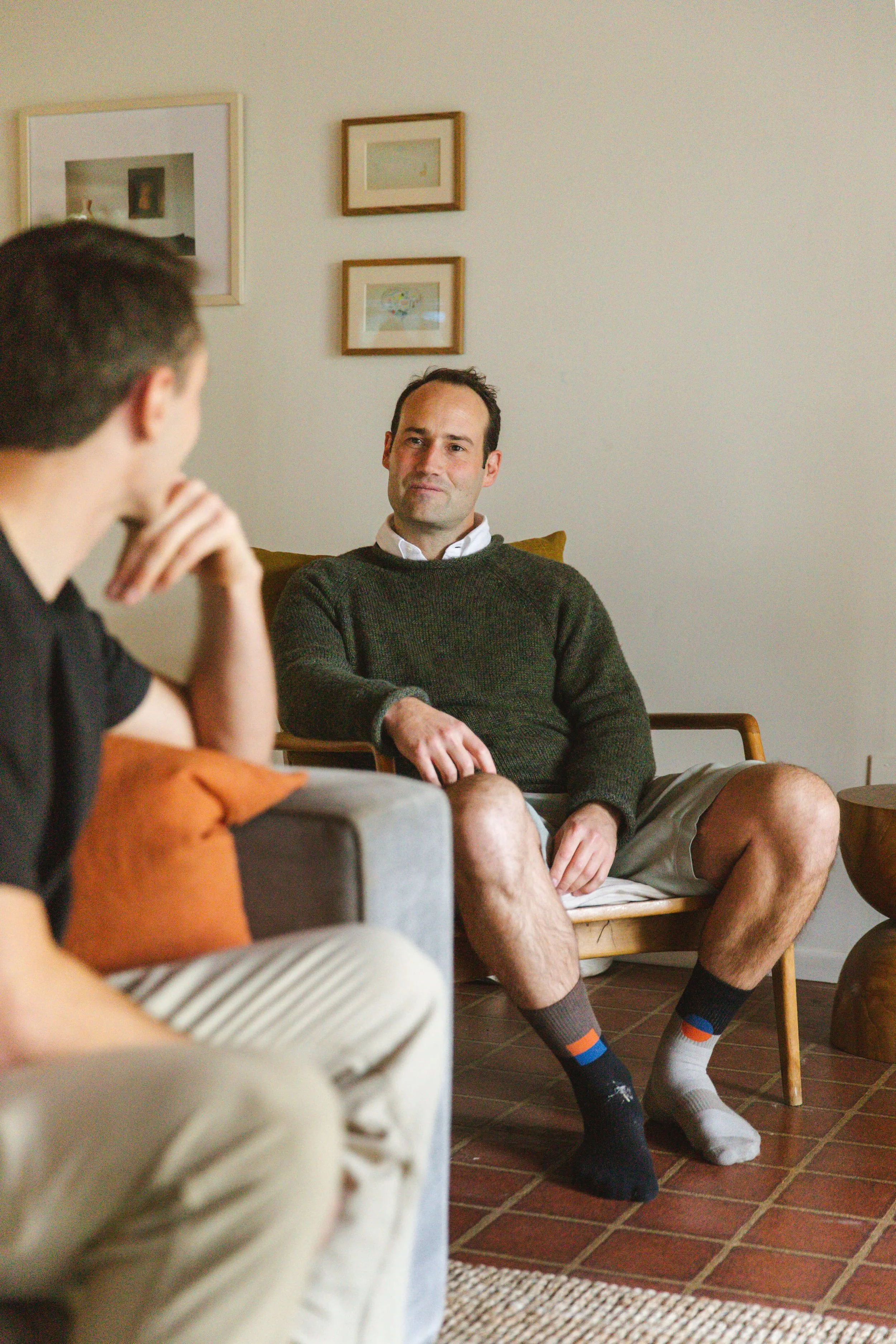 Two men sitting on a wooden chair and a gray sofa, engaging in conversation in a cozy living room with white walls and gallery wall art.