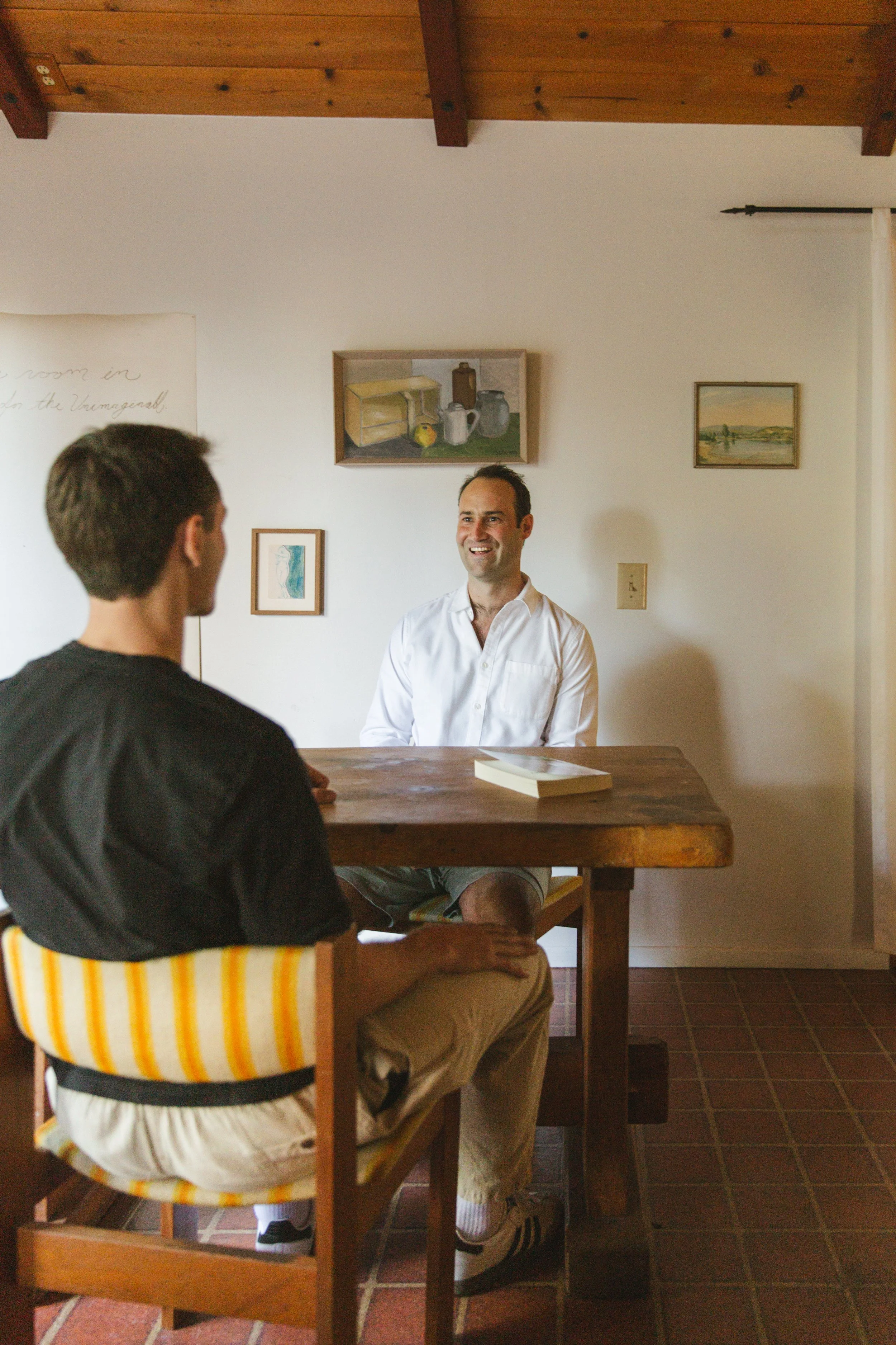 Two men having a conversation in a room with artwork on the wall. The man in the foreground has dark hair, wearing a black jacket and beige pants, sitting on a striped yellow and white chair. The man in the background has short hair, wearing a white shirt and beige shorts, sitting at a wooden table.