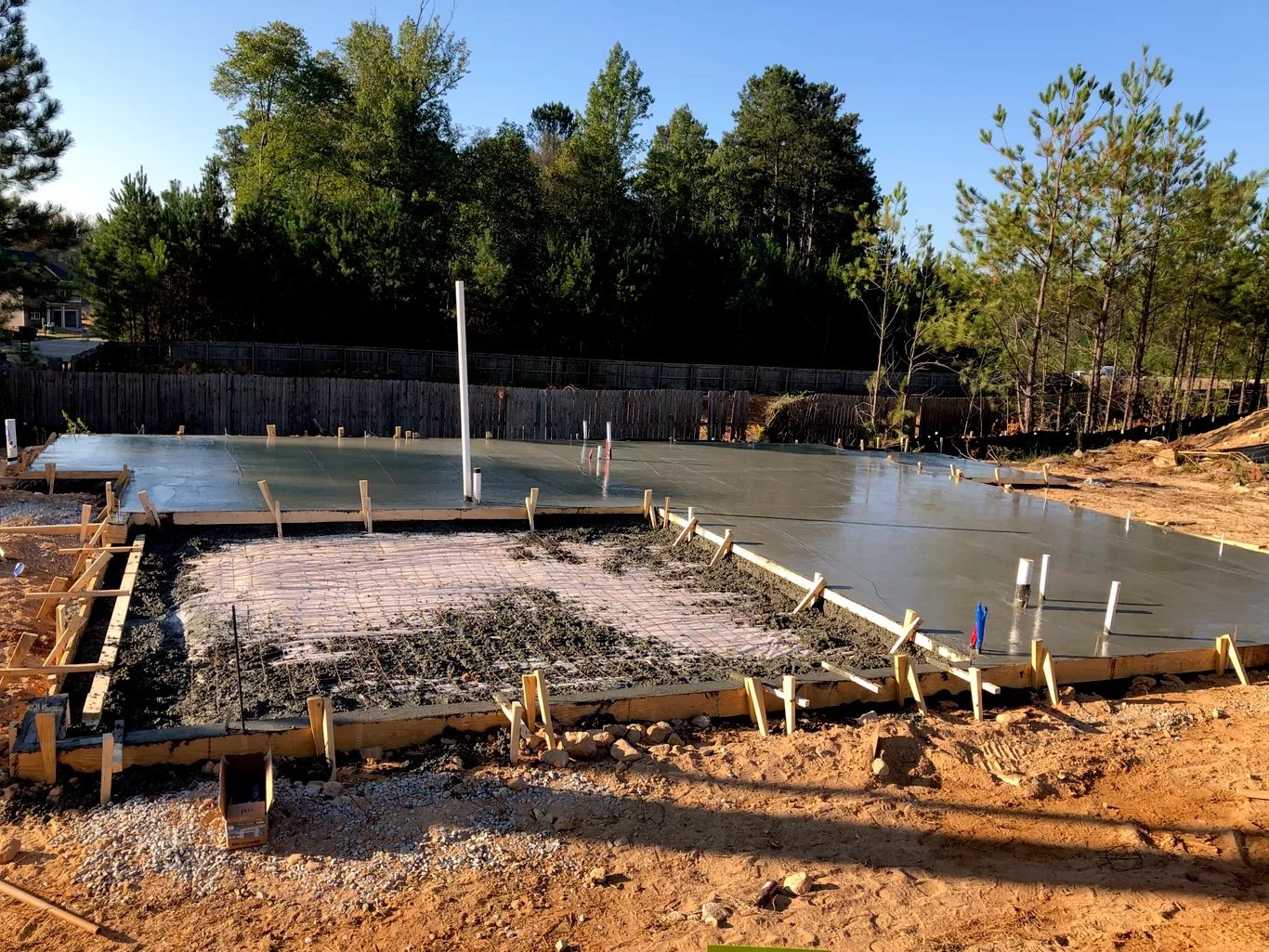 Concrete foundation with rebar and wooden framing for a building under construction, surrounded by dirt and trees in the background.