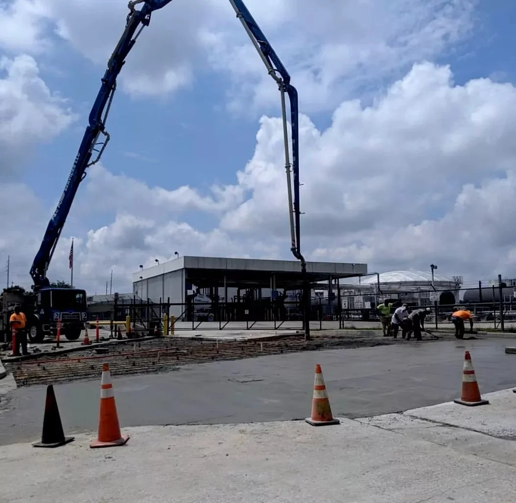 Construction workers pouring concrete on a road with a large concrete pump truck in the background and orange safety cones placed around the site.