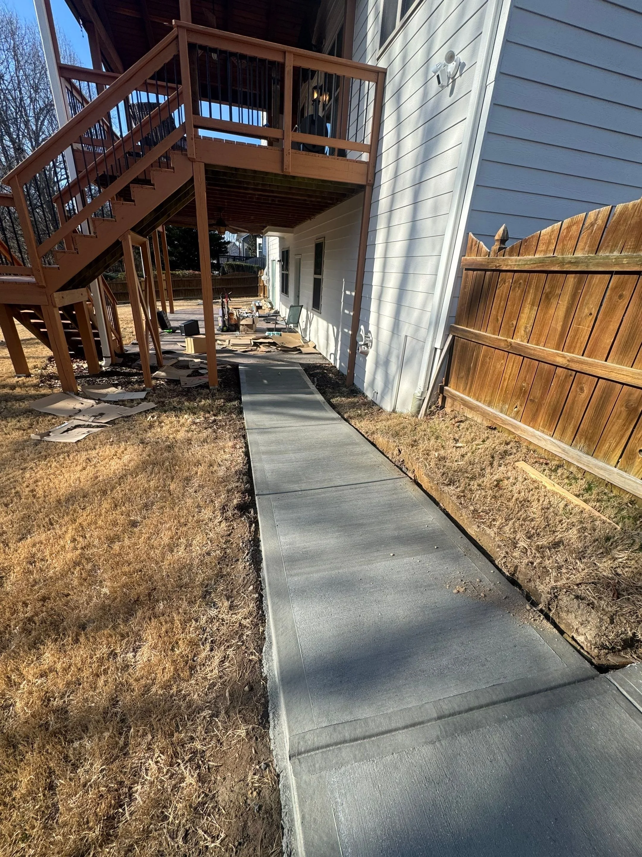 Side yard with newly poured concrete sidewalk leading to patio with wooden stairs and deck attached to white house, brown wooden fence on the right, and outdoor furniture and tools near the house.