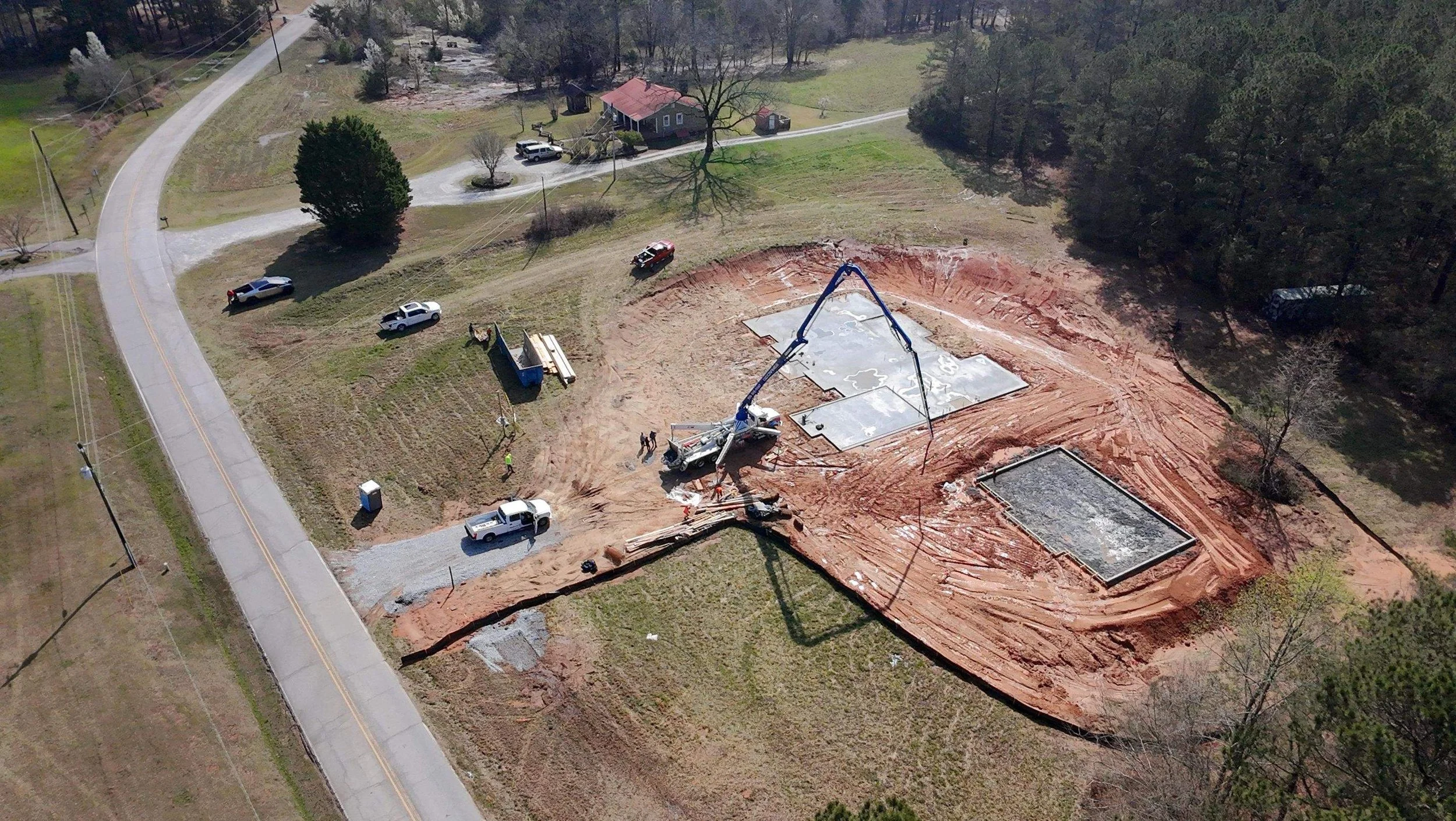 An aerial view of a construction site where foundation work is in progress. Several trucks and workers are present, and concrete slabs are being poured. The site is surrounded by grass, trees, and a curving road.