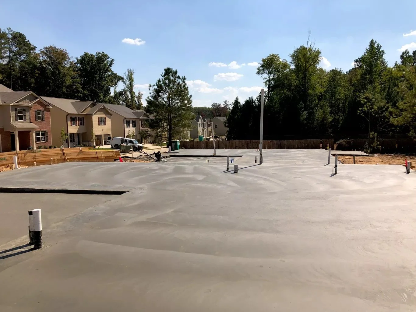 Construction site with a freshly poured concrete foundation, some pipes and poles protruding, and houses with trees in the background under a blue sky with a few clouds.