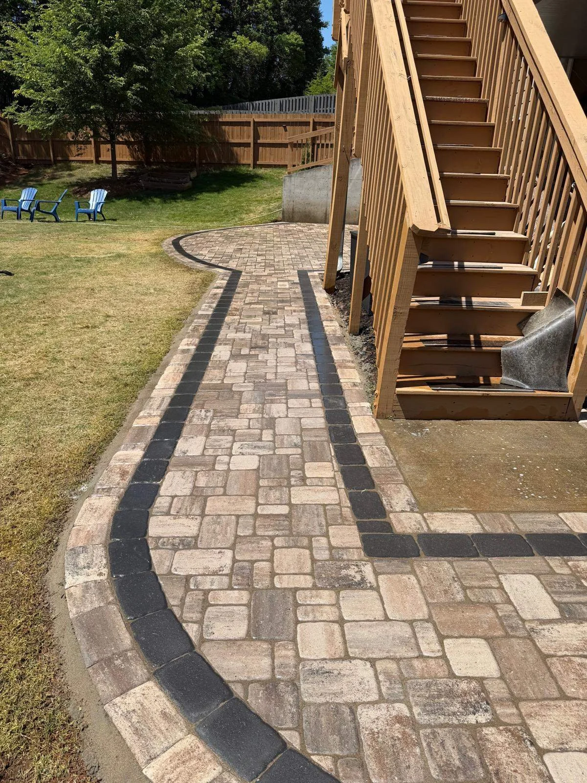 Custom stamped concrete walkway and patio area mimicking stone pavers, featuring a dark contrasting decorative border, leading to wooden deck stairs in a residential backyard.