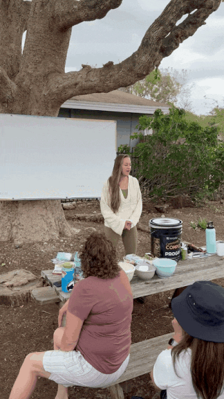 A woman in a purple shirt sitting outdoors at a picnic table, listening to a girl speaking in front of a large tree. The girl is standing near a table with food and drink items, speaking to an audience of two women. There is a home and greenery in the background.