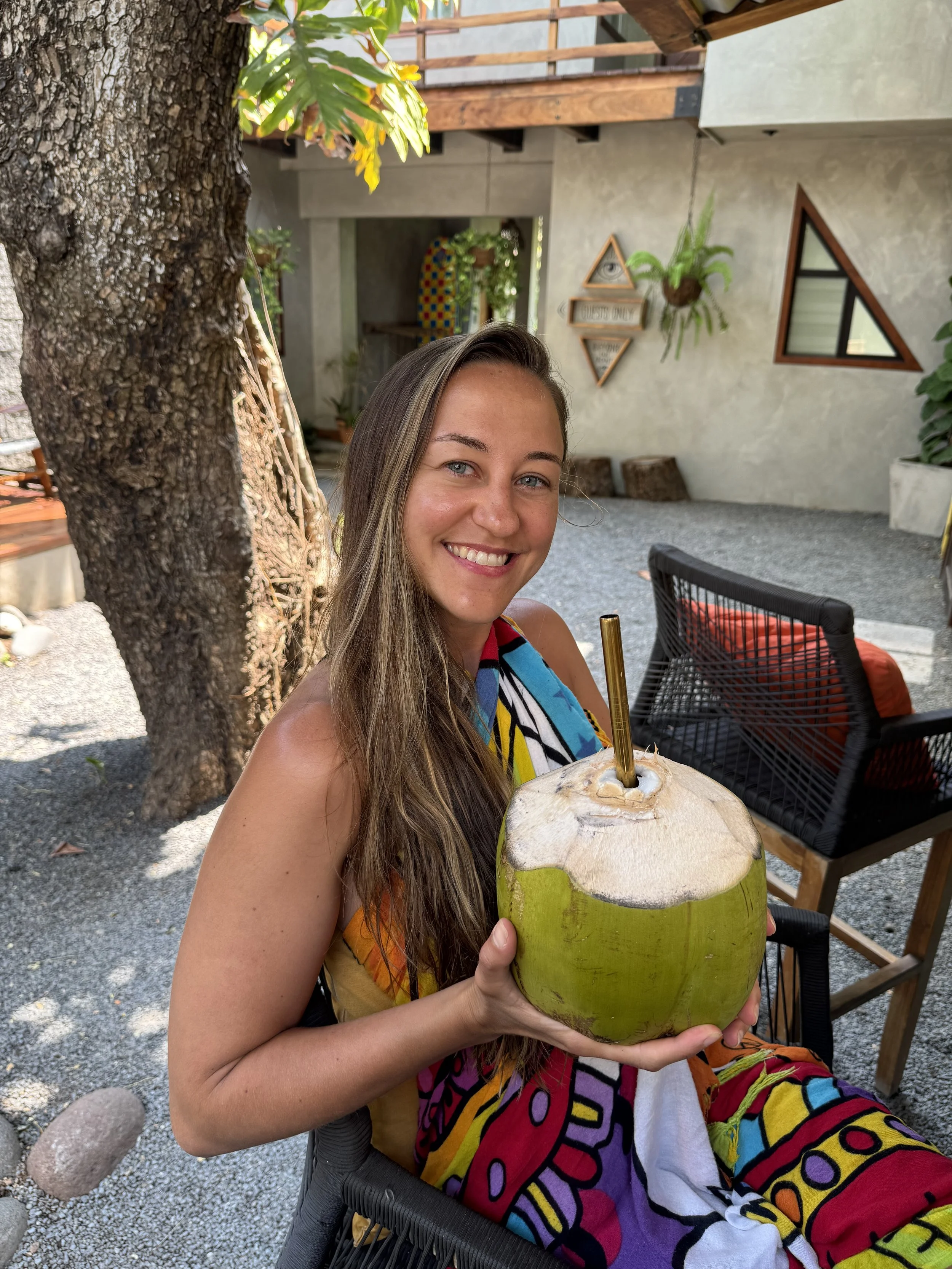 A woman smiling and holding a fresh opened green coconut with a straw, sitting outdoors in a patio area with trees, plants, wooden furniture, and colorful decor.
