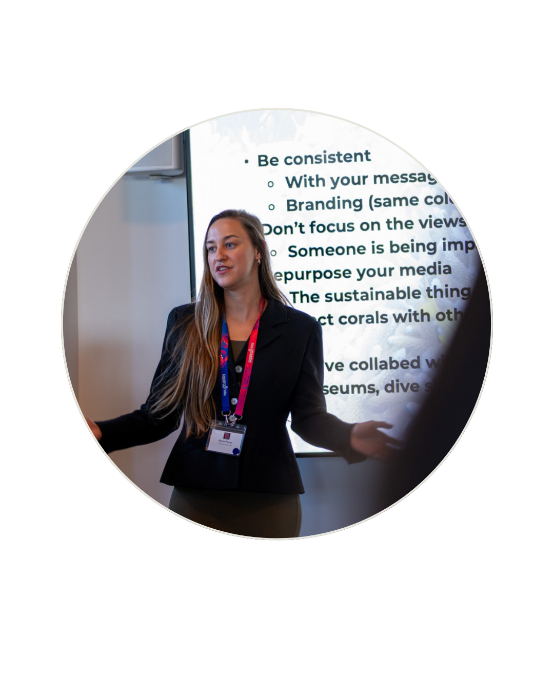 A woman in a black blazer and a name badge giving a presentation in front of a screen with text on it.