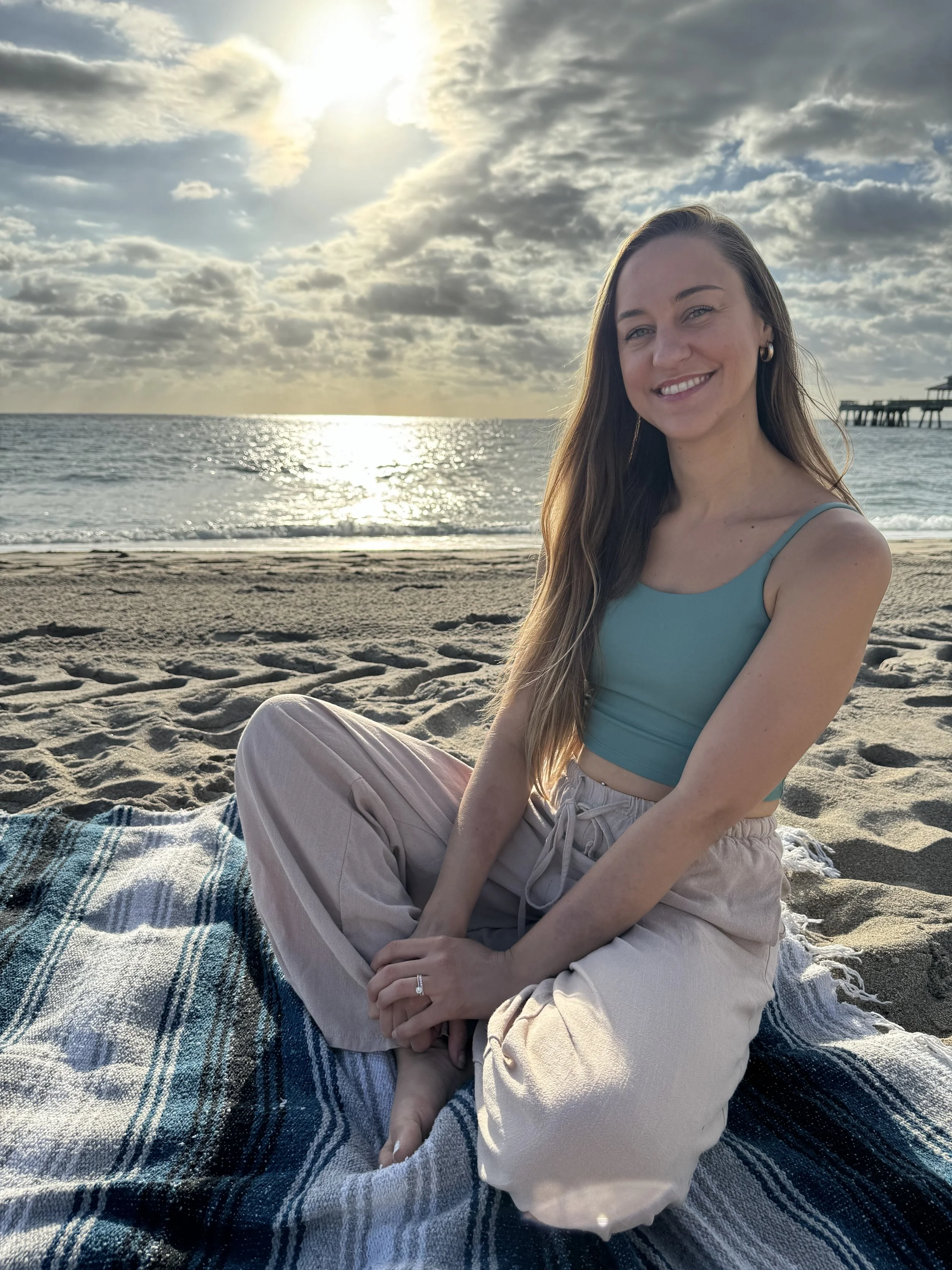A woman sitting on a beach on a blanket, smiling, with the ocean and cloudy sky in the background.