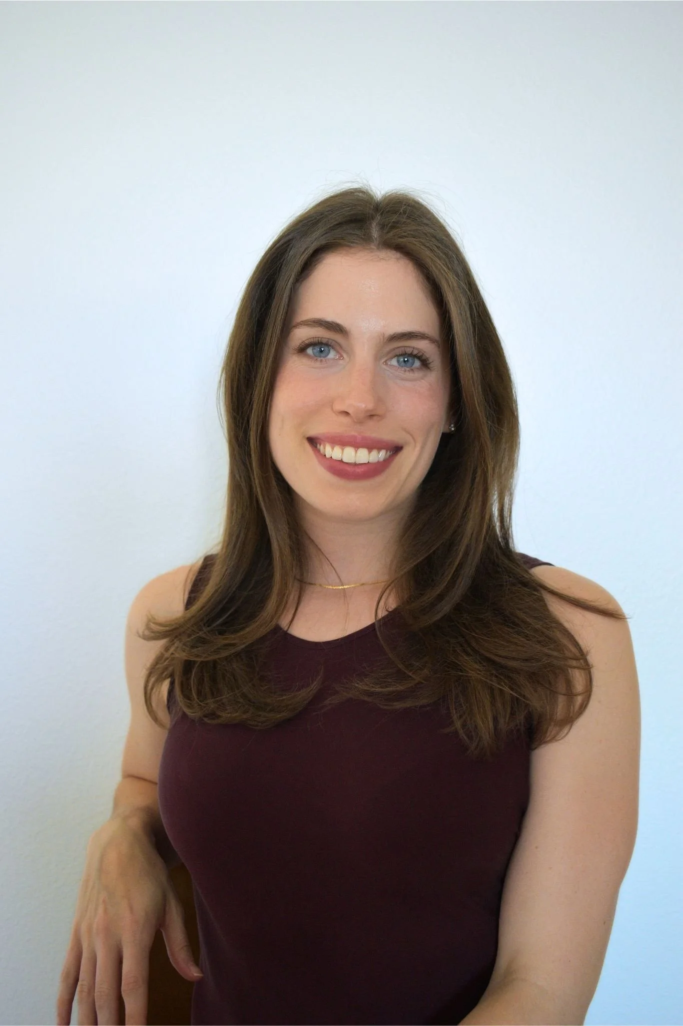 A woman smiling with long brown hair, wearing a sleeveless maroon top, and a gold necklace, against a plain white wall.