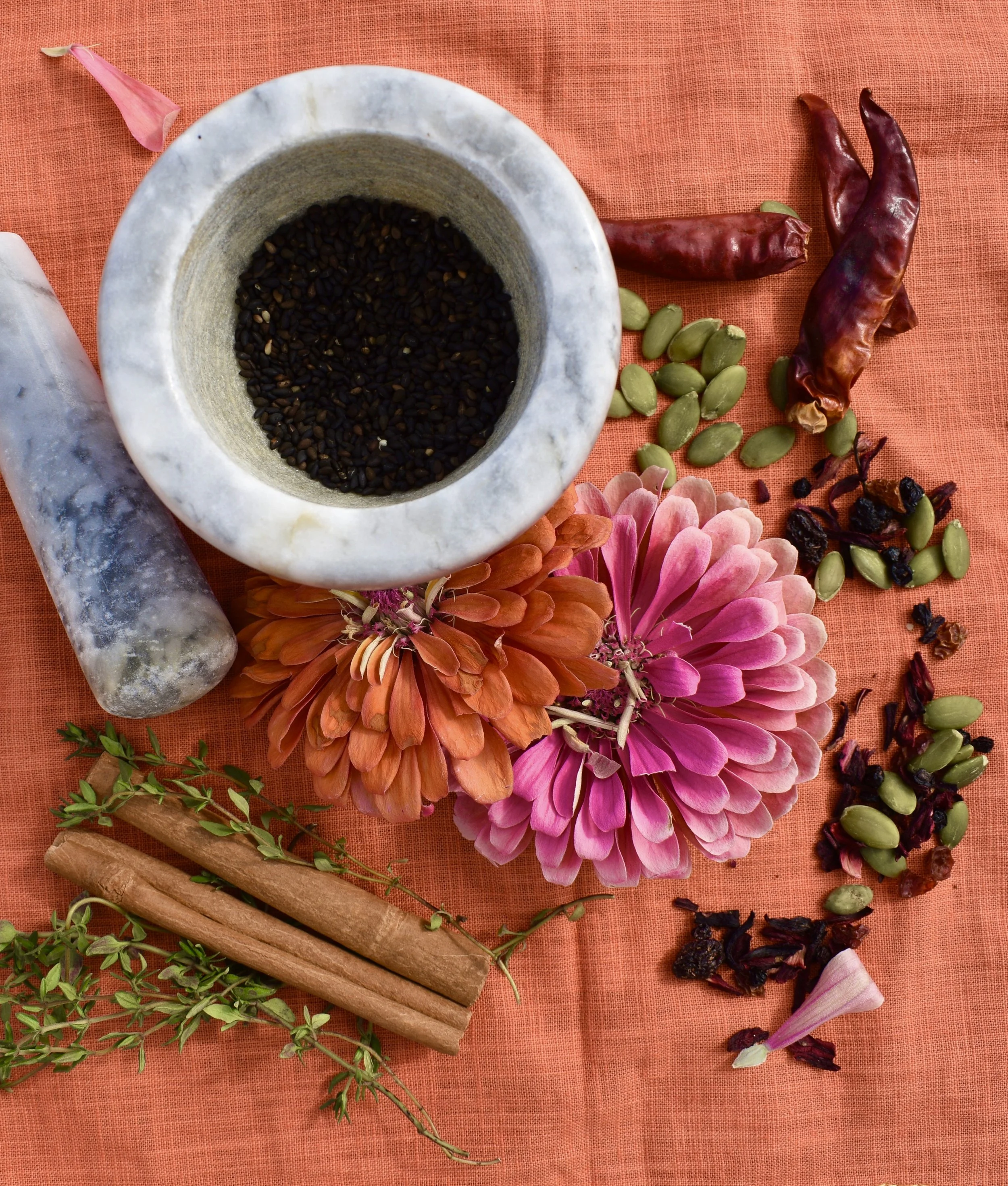 A marble mortar and pestle containing black sesame seeds on an orange fabric surface, surrounded by dried chili peppers, green pumpkin seeds, pink and orange flowers, cinnamon sticks, fresh thyme, and dried rose petals.