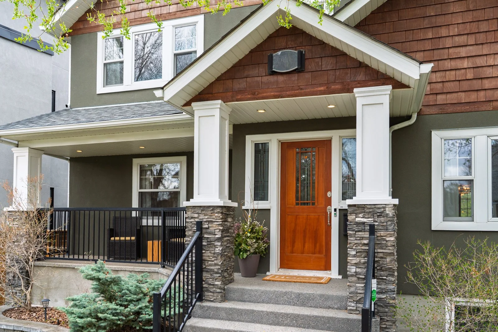 Front exterior of a house with a wooden front door, stone pillars, and a small porch with black railings. The house has green and brown siding, with a gabled roof, and is surrounded by trees and shrubs.