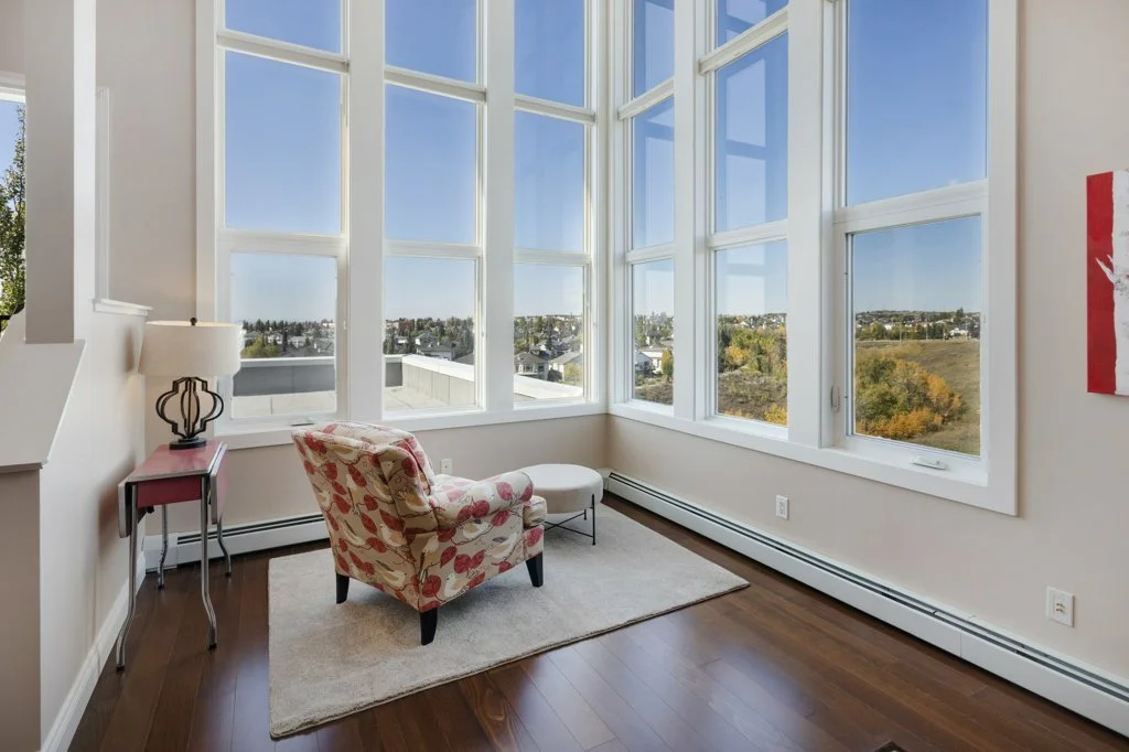 A bright corner of a room with large windows showcasing a landscape with trees and houses, a patterned armchair, a small pink side table with a lamp, and a white rug on wooden flooring.