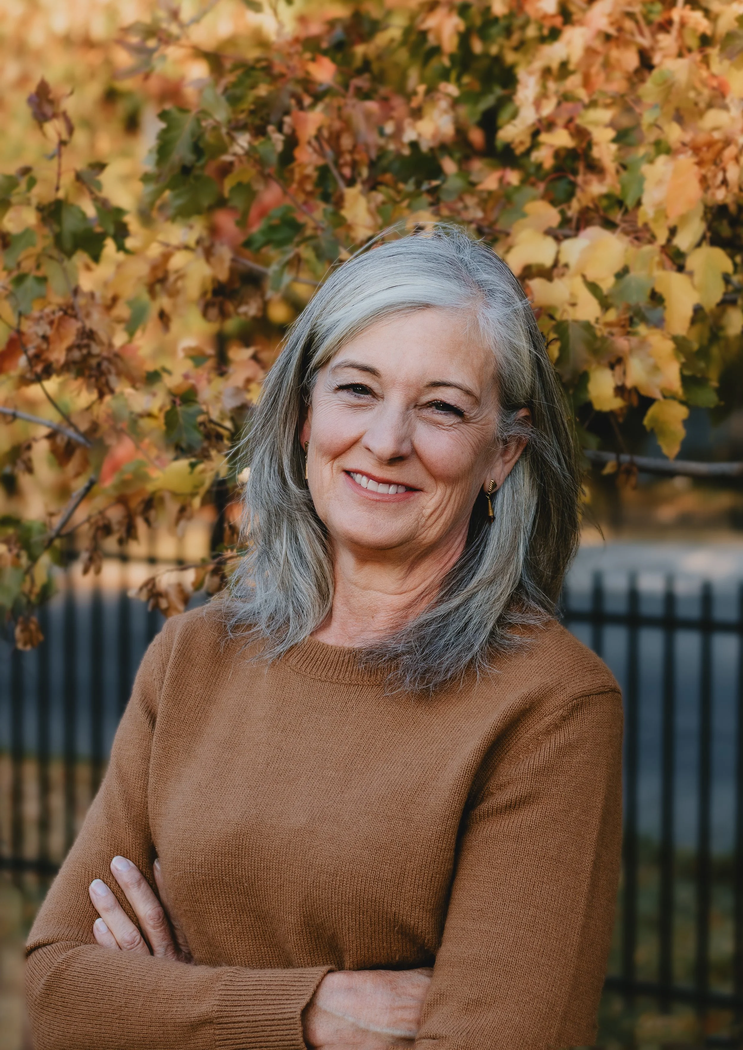 Michele Gole, Exp realty in a tan sweater standing outdoors with autumn leaves in the background.