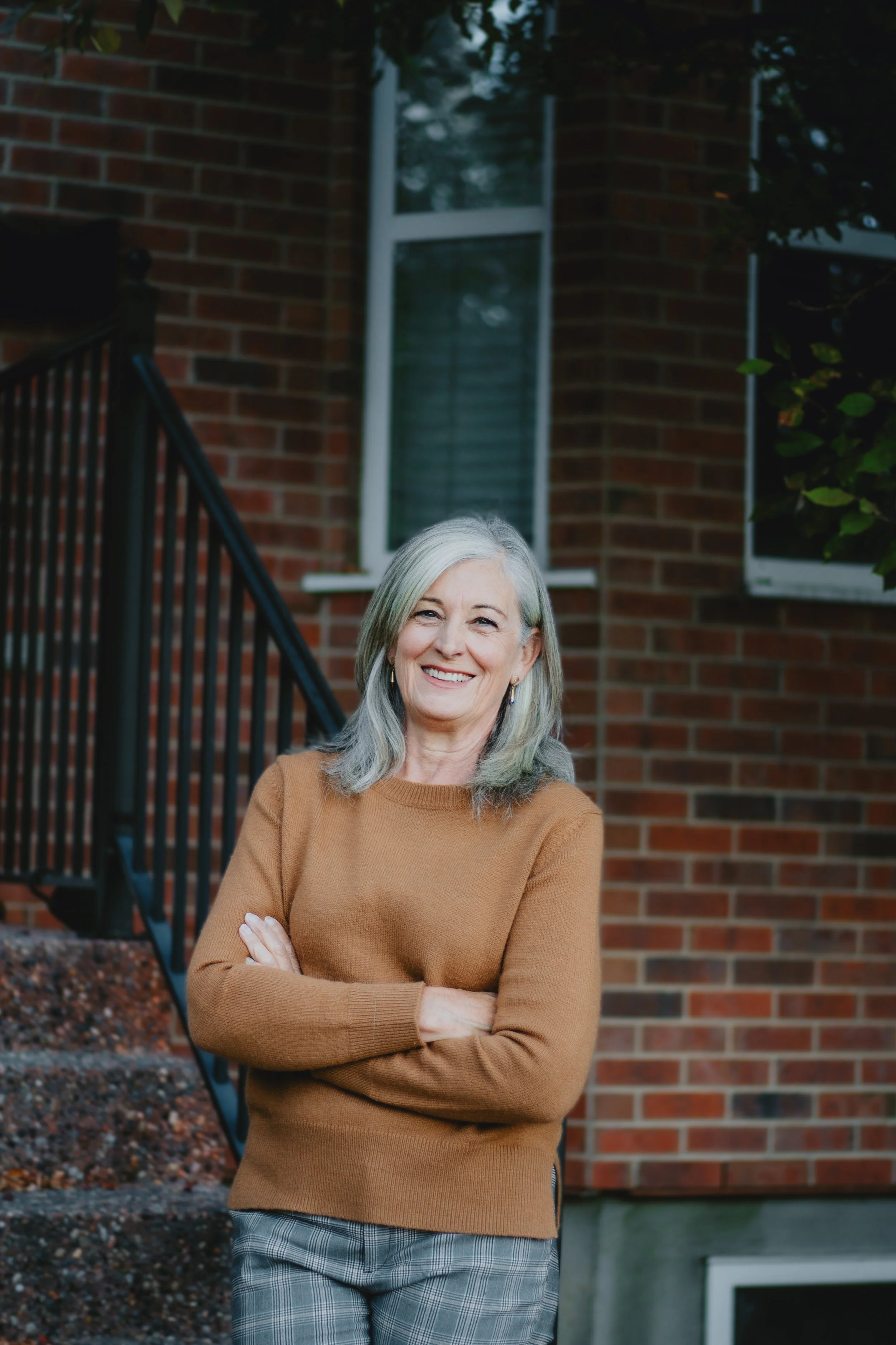 Michele Gole, Exp realty, wearing a brown sweater and gray plaid pants, standing with arms crossed outside a brick house with windows and stairs, during daytime.
