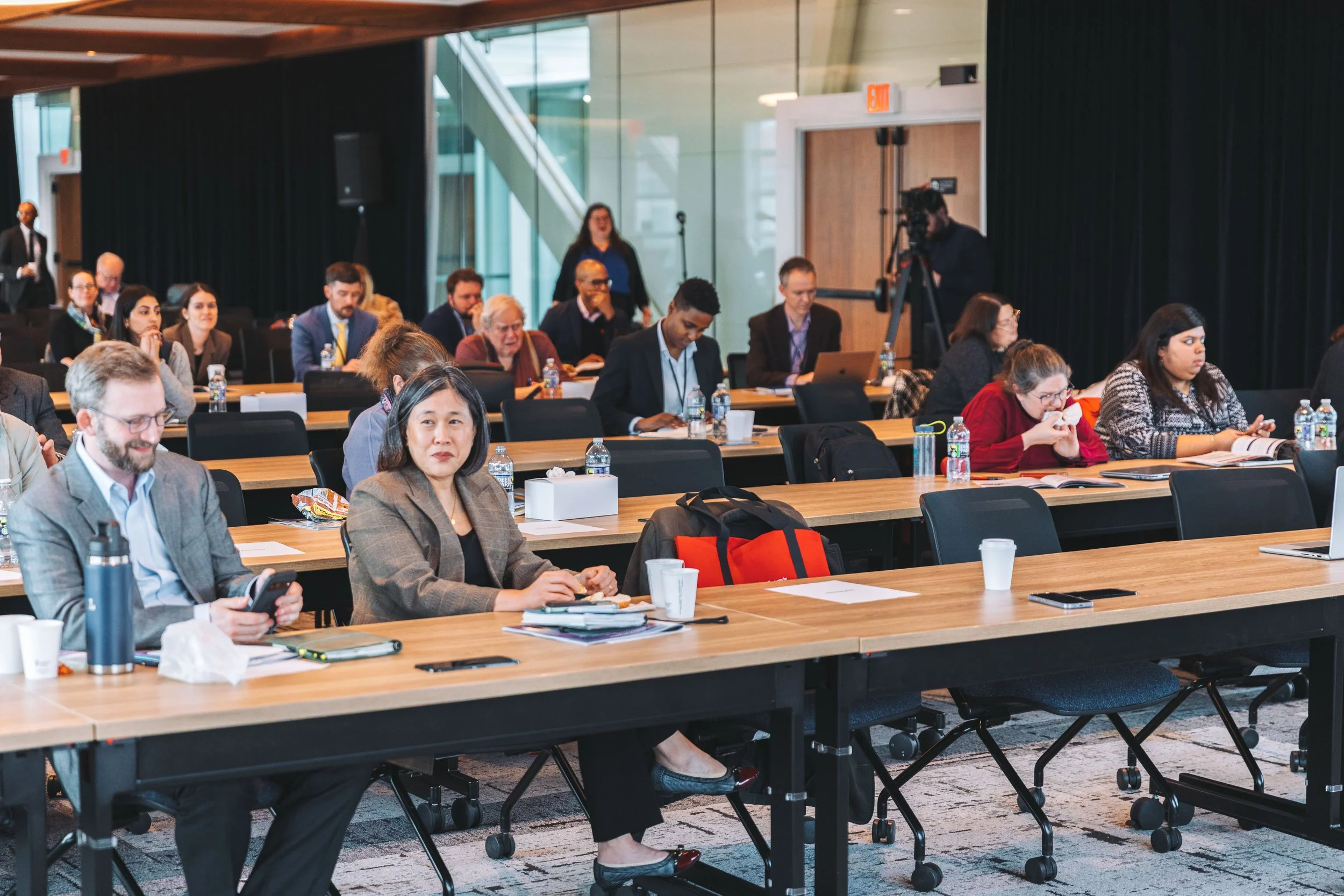Attendees seated at long tables in a conference room, some taking notes or using electronic devices, with a person operating a camera in the background.