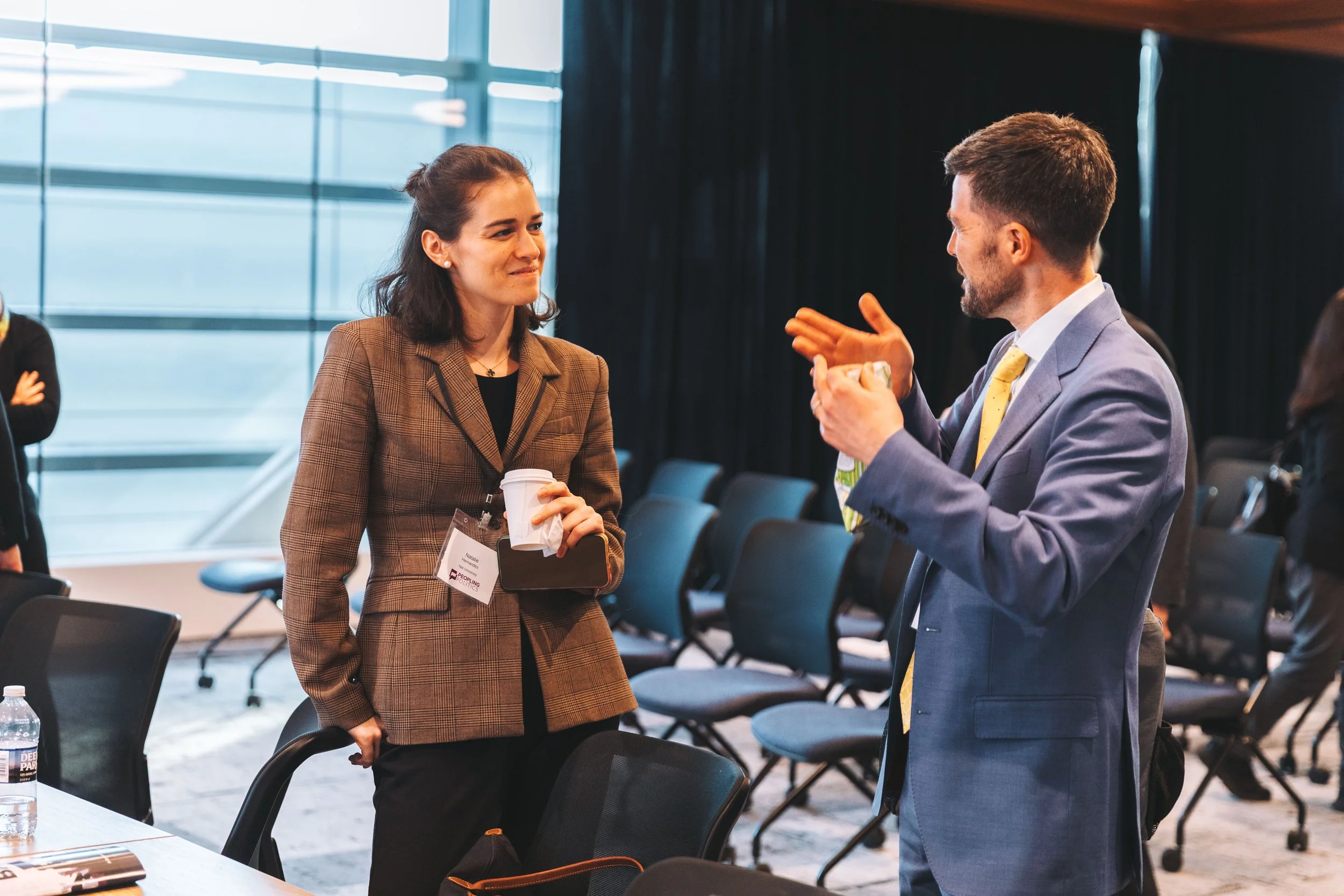 A man and woman are engaged in conversation during a conference or business event. The woman is holding a coffee cup, and both are smiling as they talk. The setting is a modern conference room with large windows and empty chairs in the background.
