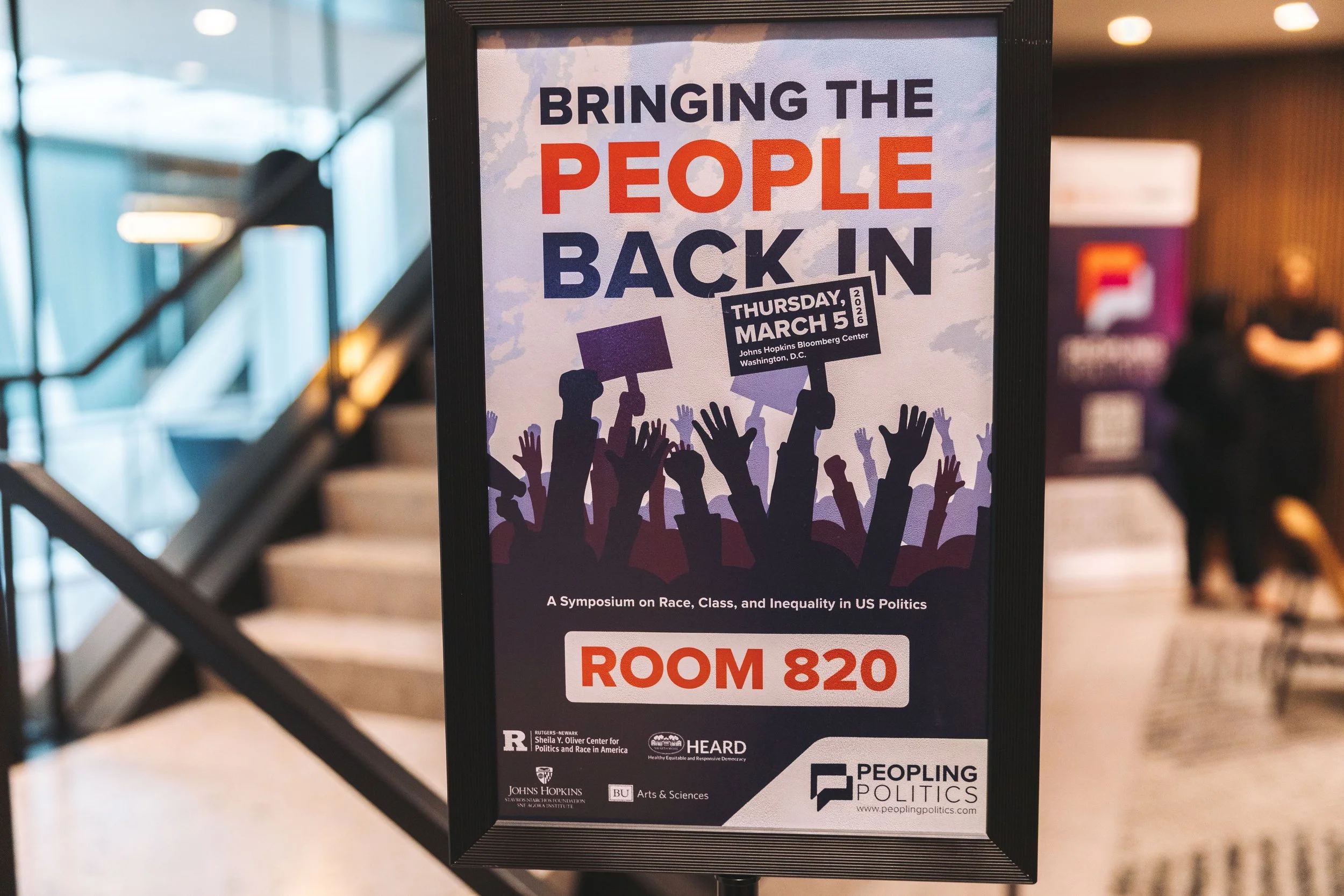 Sign advertising a symposium titled 'Bringing the People Back In' scheduled for Thursday, March 5, 2020, at Johns Hopkins Bloomberg Center, Washington D.C., with room number 820. The sign features silhouettes of a crowd holding signs and raising thei