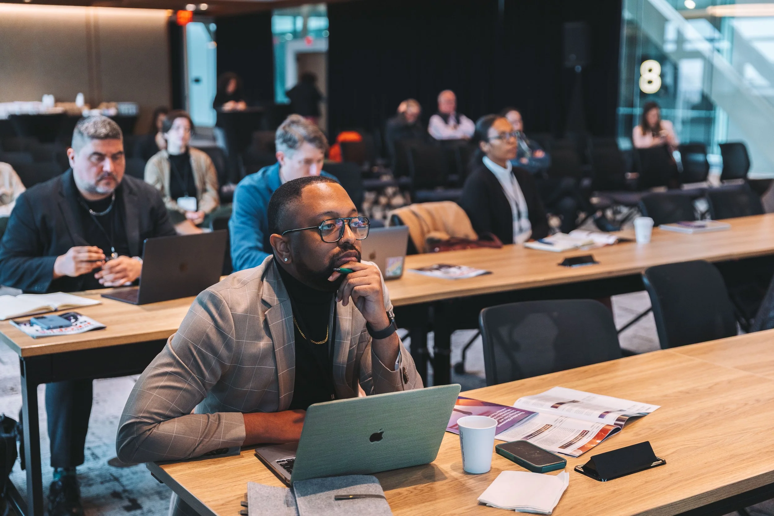 A group of diverse people seated at long conference tables, listening to a presentation in a modern meeting room.