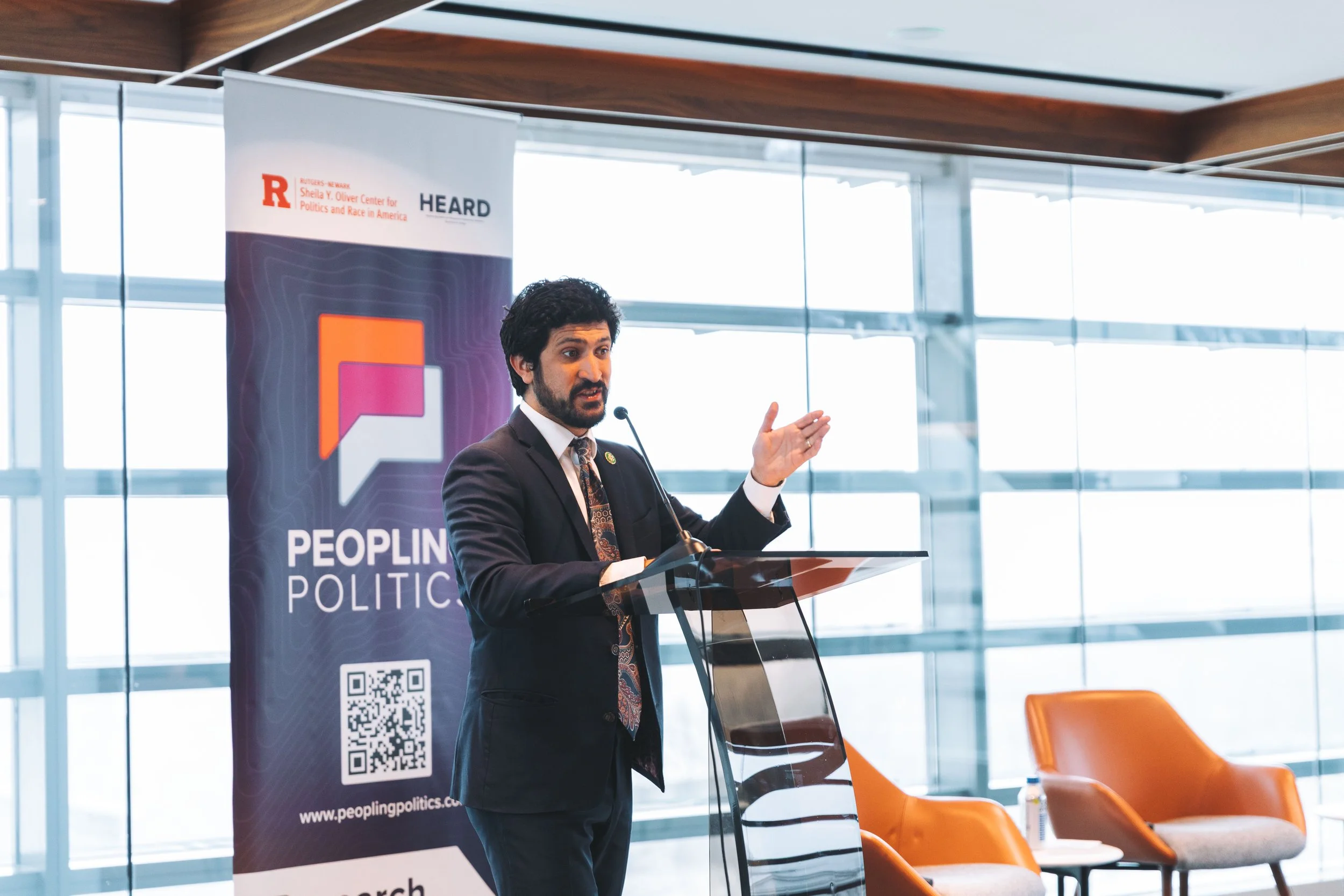 A man in a black suit and patterned tie is giving a speech at a podium with a microphone. Behind him, there is a vertical banner with the words "Peoplings Politics" and a QR code. The background consists of large glass windows and two orange chairs w