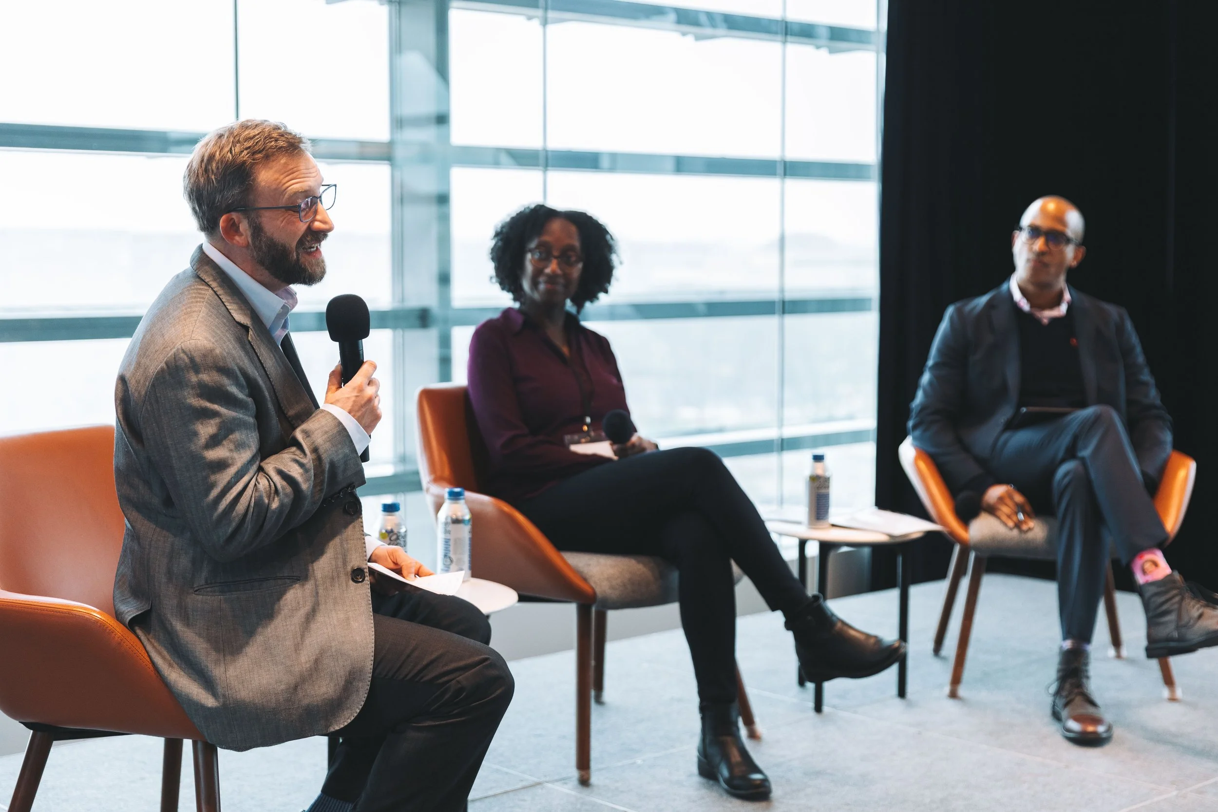 A panel discussion with three people seated with microphones, water bottles, and notes, in a modern office setting with large windows.