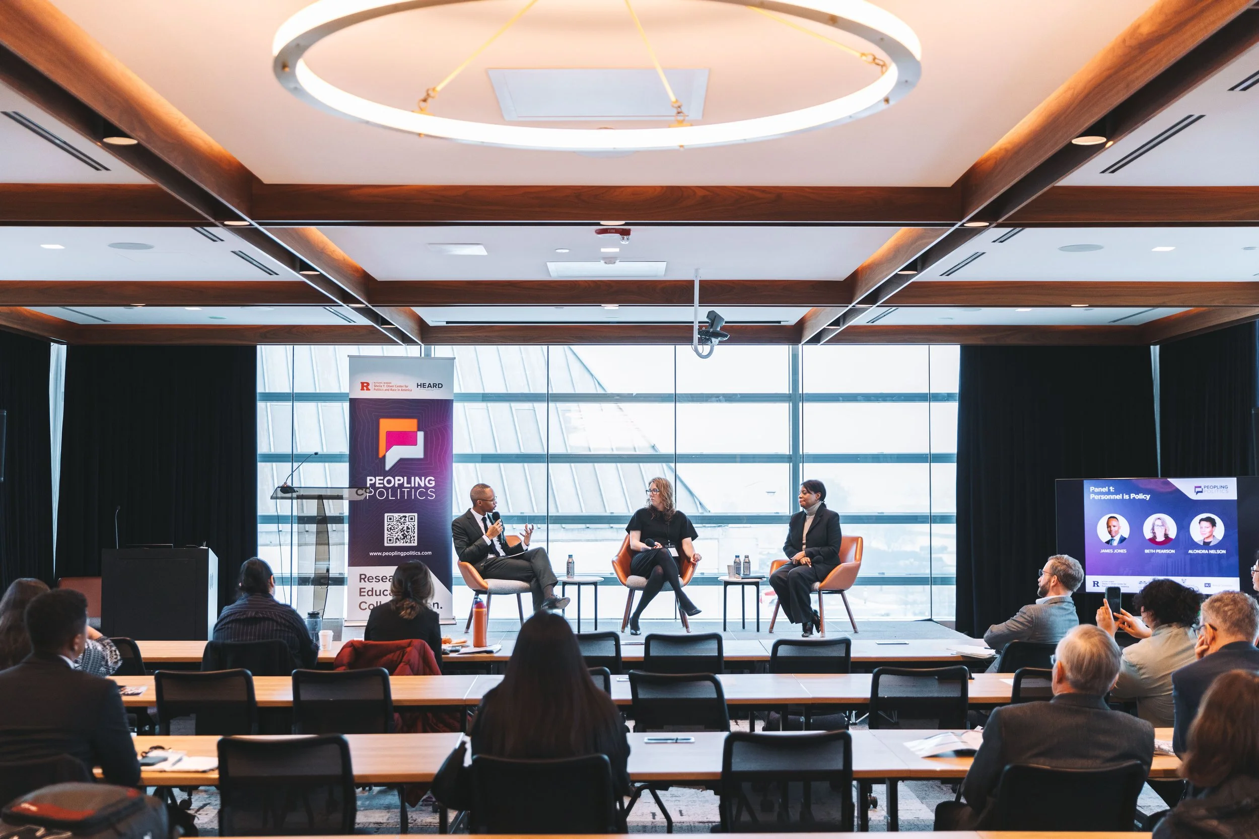 A panel discussion at a conference with three speakers on stage. One man is speaking, with two women listening, in front of large windows showing a cityscape. Audience members are seated facing the stage, some taking photos.