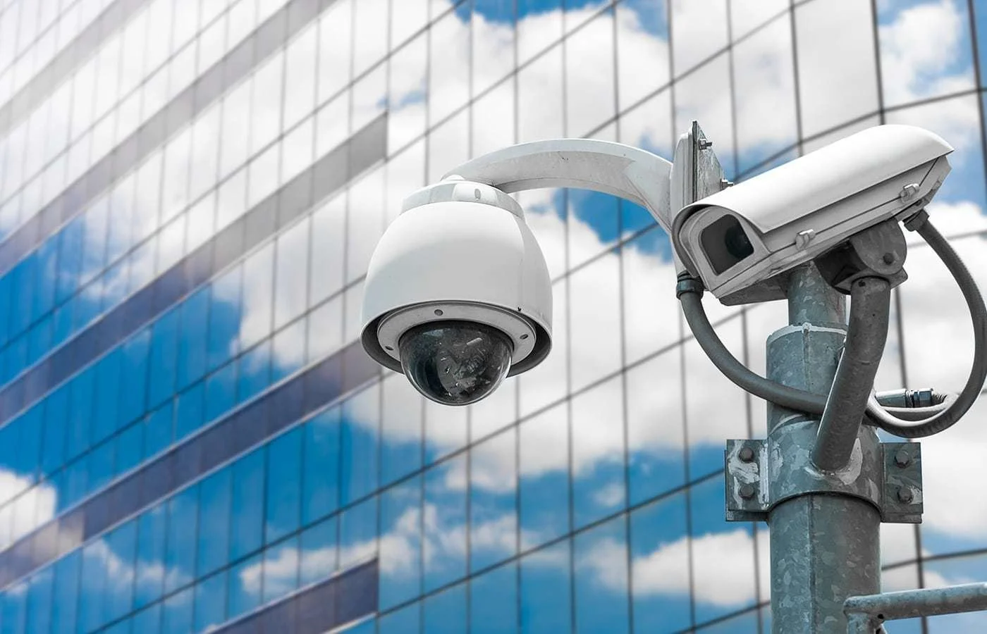 Security camera mounted on a pole in front of a modern glass building with reflected blue sky and clouds.