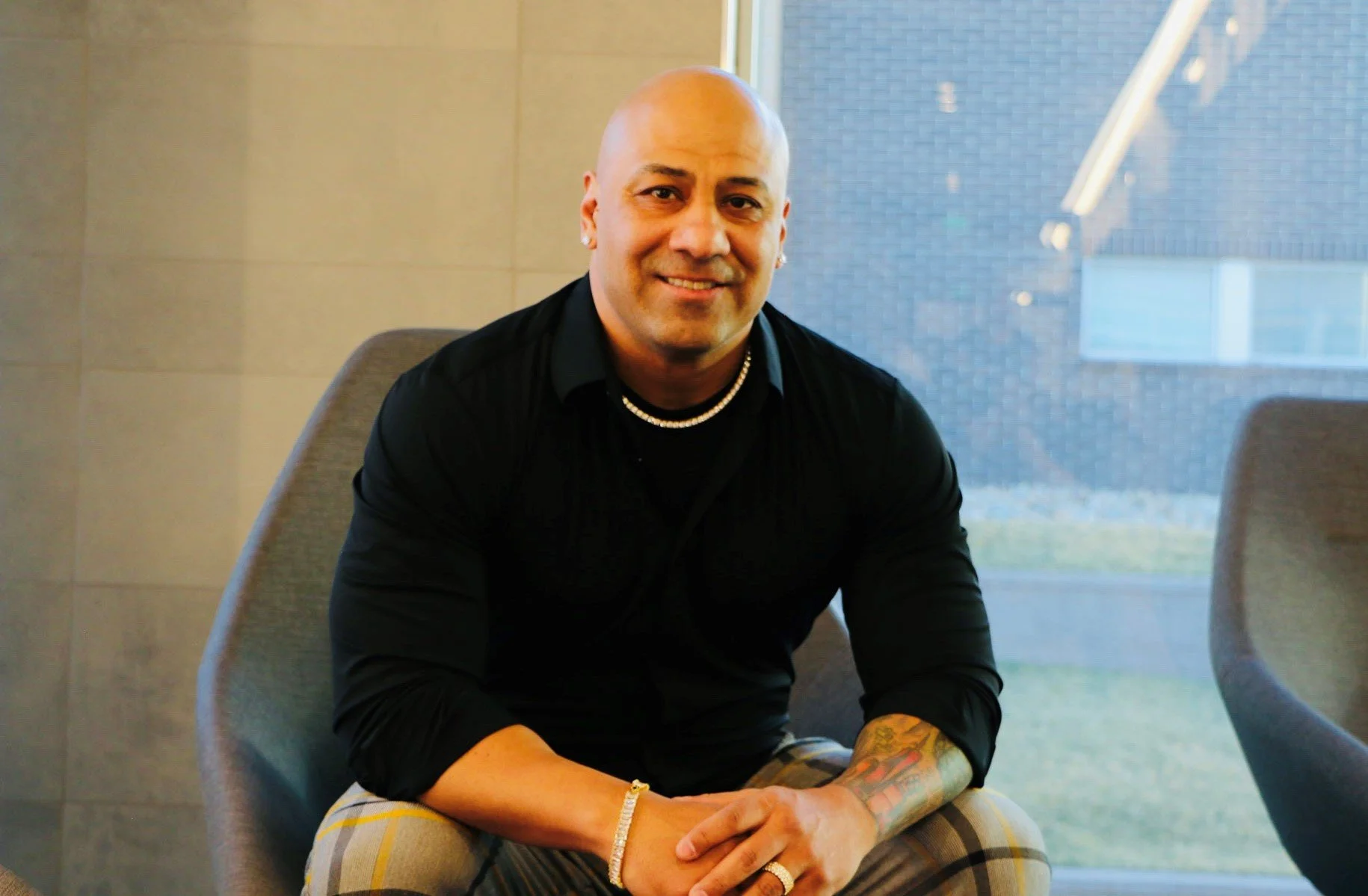 A smiling man with a shaved head sitting in a modern indoor space, wearing a black shirt, checkered pants, a pearl necklace, and jewelry, with a window in the background showing a building and grass outside.
