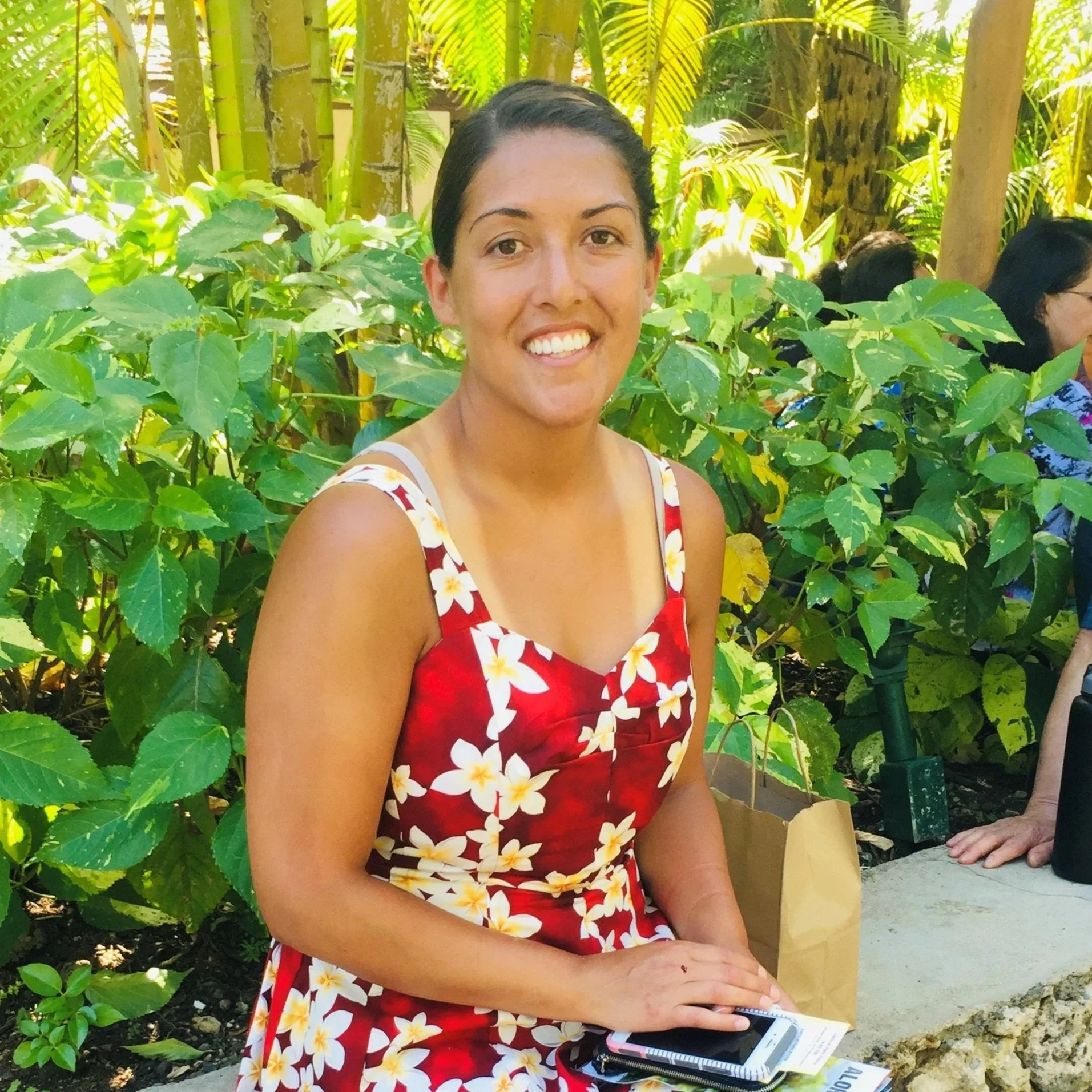 A smiling woman with dark hair in a floral red and white dress sitting outdoors among green plants with bamboo in the background.