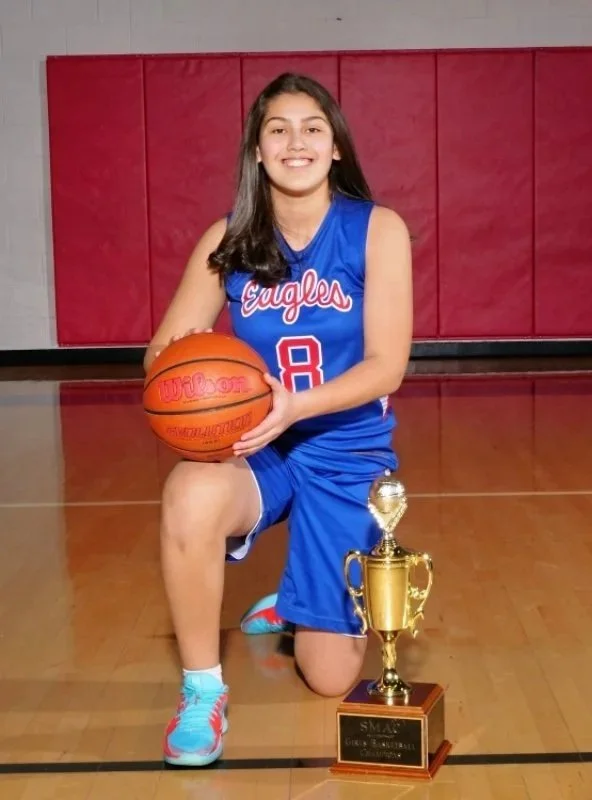 A young girl in a blue basketball uniform with the number 8, sitting on one knee on a basketball court, holding a basketball, and smiling. There is a large trophy placed in front of her on the floor. The background shows red padding on the wall of the gymnasium.