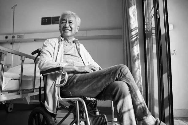 An elderly person sitting in a wheelchair in a hospital room, smiling, near a window with sunlight.