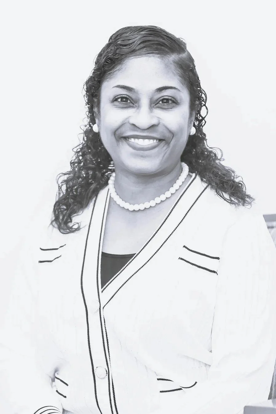 A black-and-white portrait of a smiling woman with curly hair wearing a white blazer with black trim, a black top, a pearl necklace, and pearl earrings.