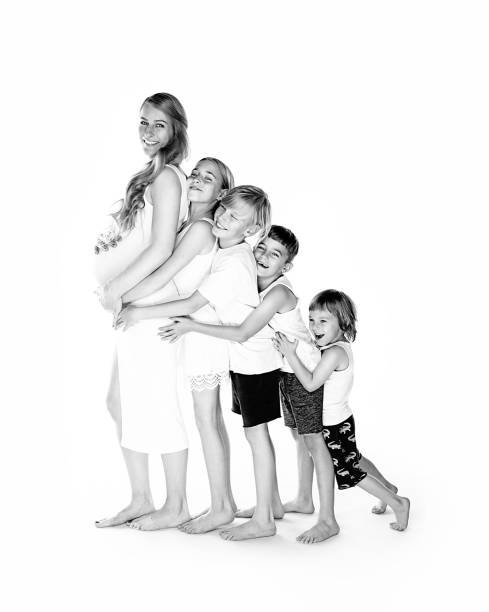 Black and white photo of a woman with four children hugging her in a playful pose, all smiling and standing against a plain background.