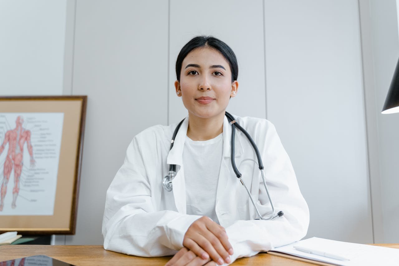 A female doctor sitting at a desk with a stethoscope around her neck, in a medical office with a anatomy chart on the wall.