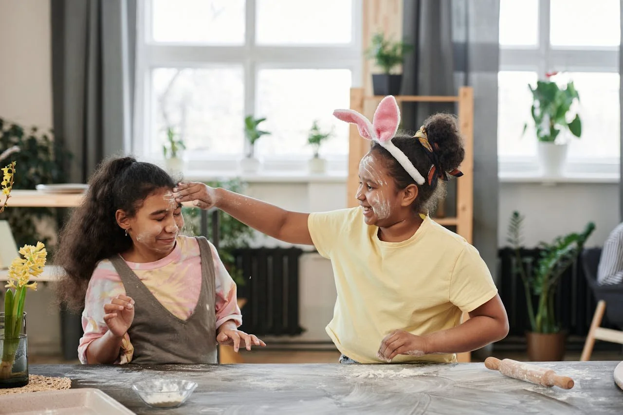 Two girls in a bright kitchen. One girl is playfully dabbing flour on the other girl's forehead while both are smiling. One girl is wearing bunny ears.