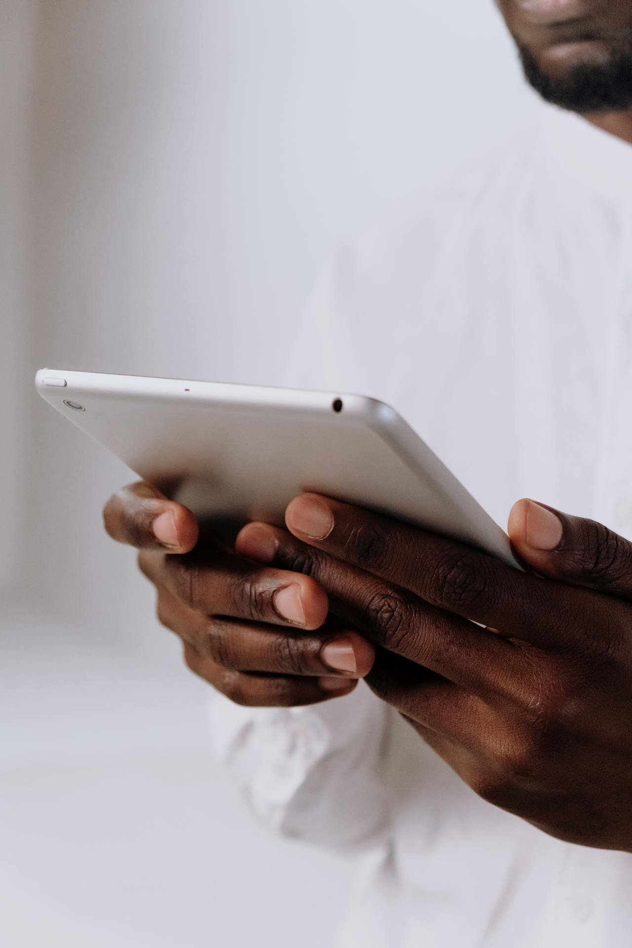 Close-up of a person with dark skin holding a white tablet with both hands, wearing a white shirt, against a plain background.