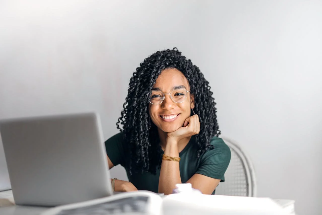 A woman with curly black hair, wearing glasses and a green shirt, smiling at the camera while sitting at a desk with a laptop.