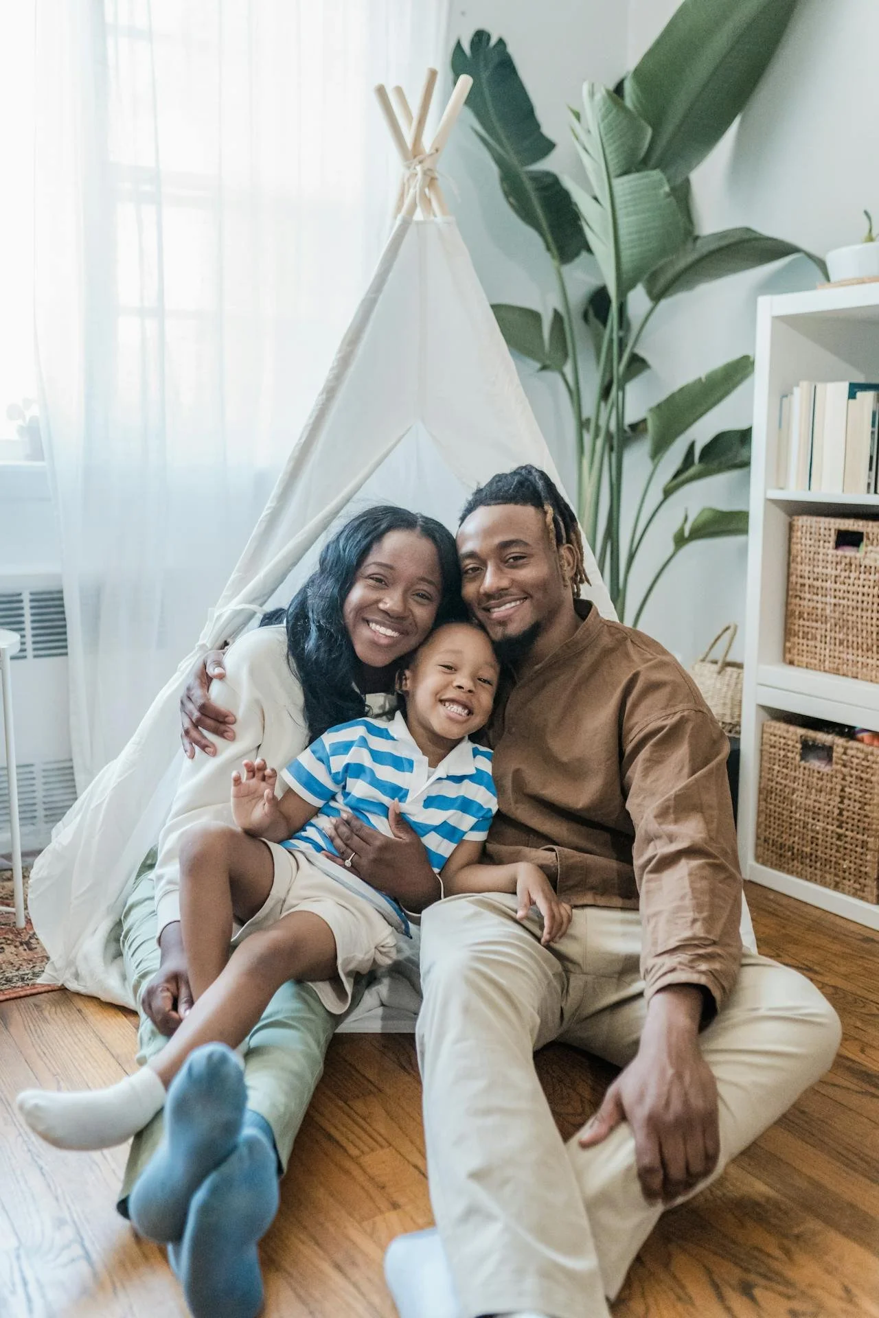 Family of three sitting on the floor inside a room with a teepee tent, large green houseplants, and a white bookshelf with baskets and books.