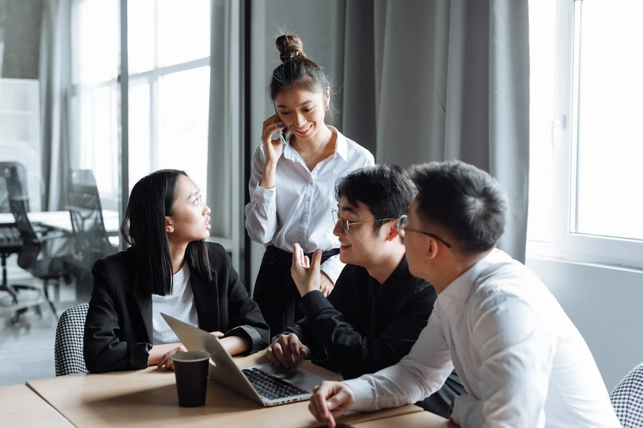 Four young professionals in an office, engaged in a discussion around a laptop, with one woman standing and talking on the phone.