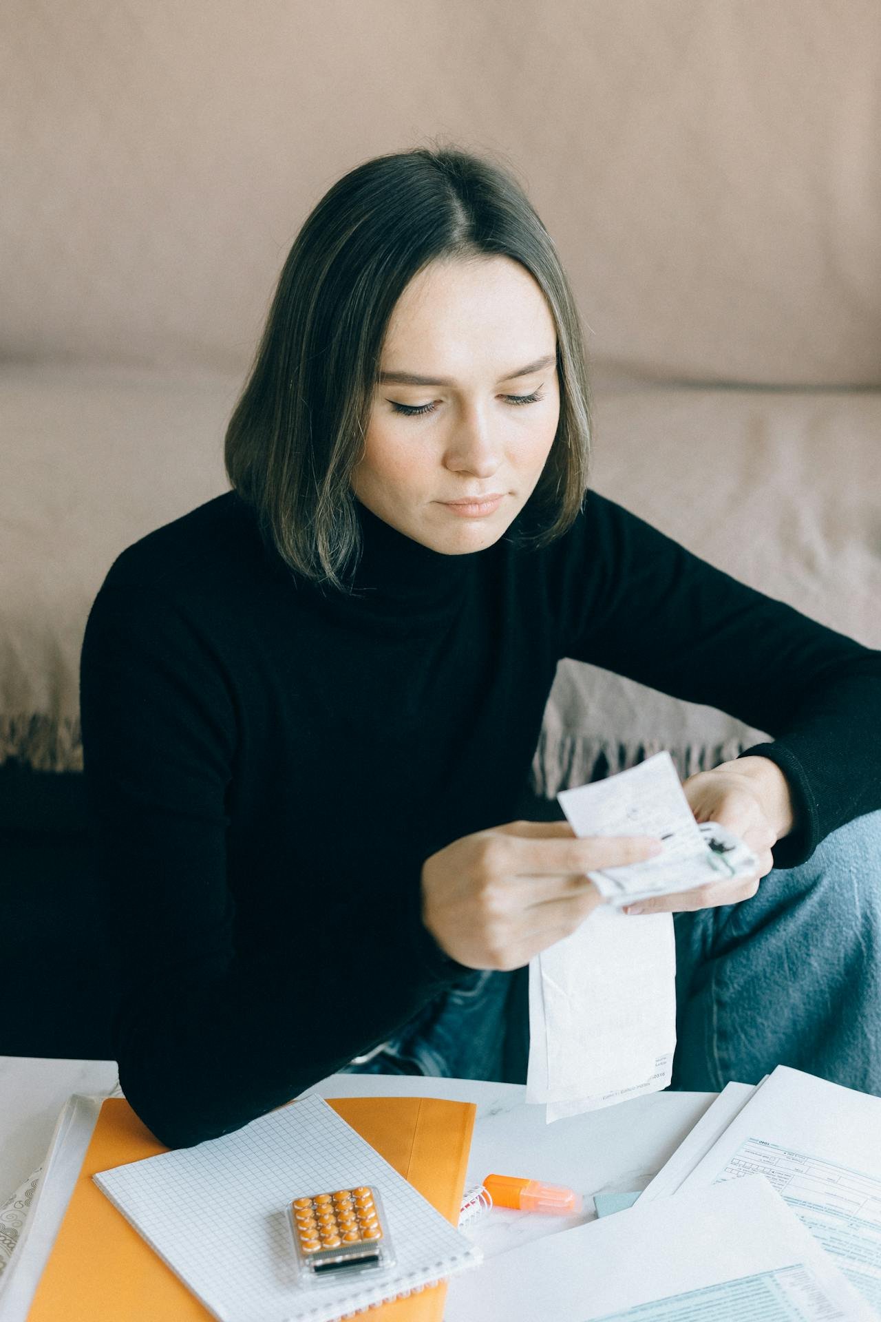 A woman with shoulder-length hair wearing a black turtleneck sitting at a table with papers, a notebook, a calculator, and a highlighter, looking at a receipt in her hand.