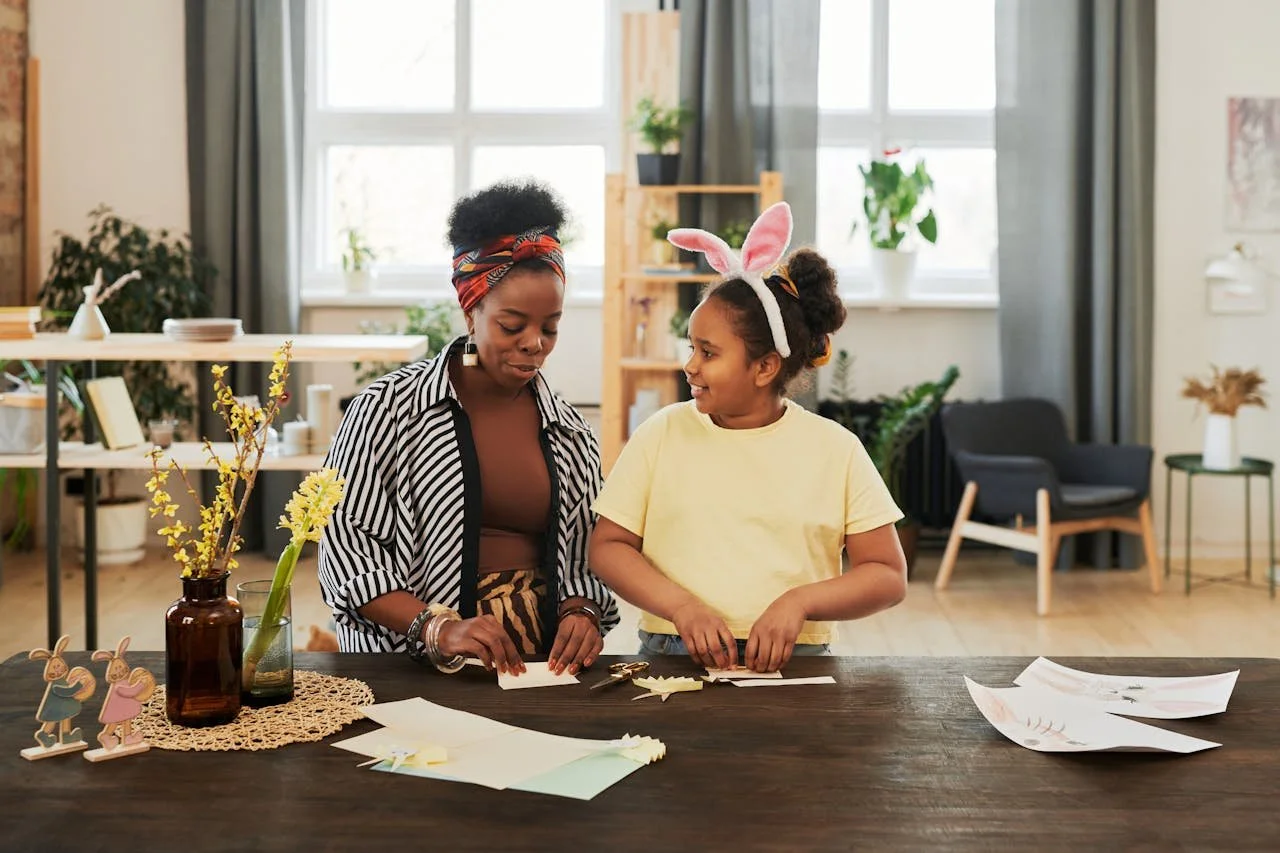 A woman and a girl with bunny ears in a cozy, well-lit living room. They are organizing Easter decorations on a wooden table, which holds paper cutouts, a vase with yellow flowers, and bunny figurines.