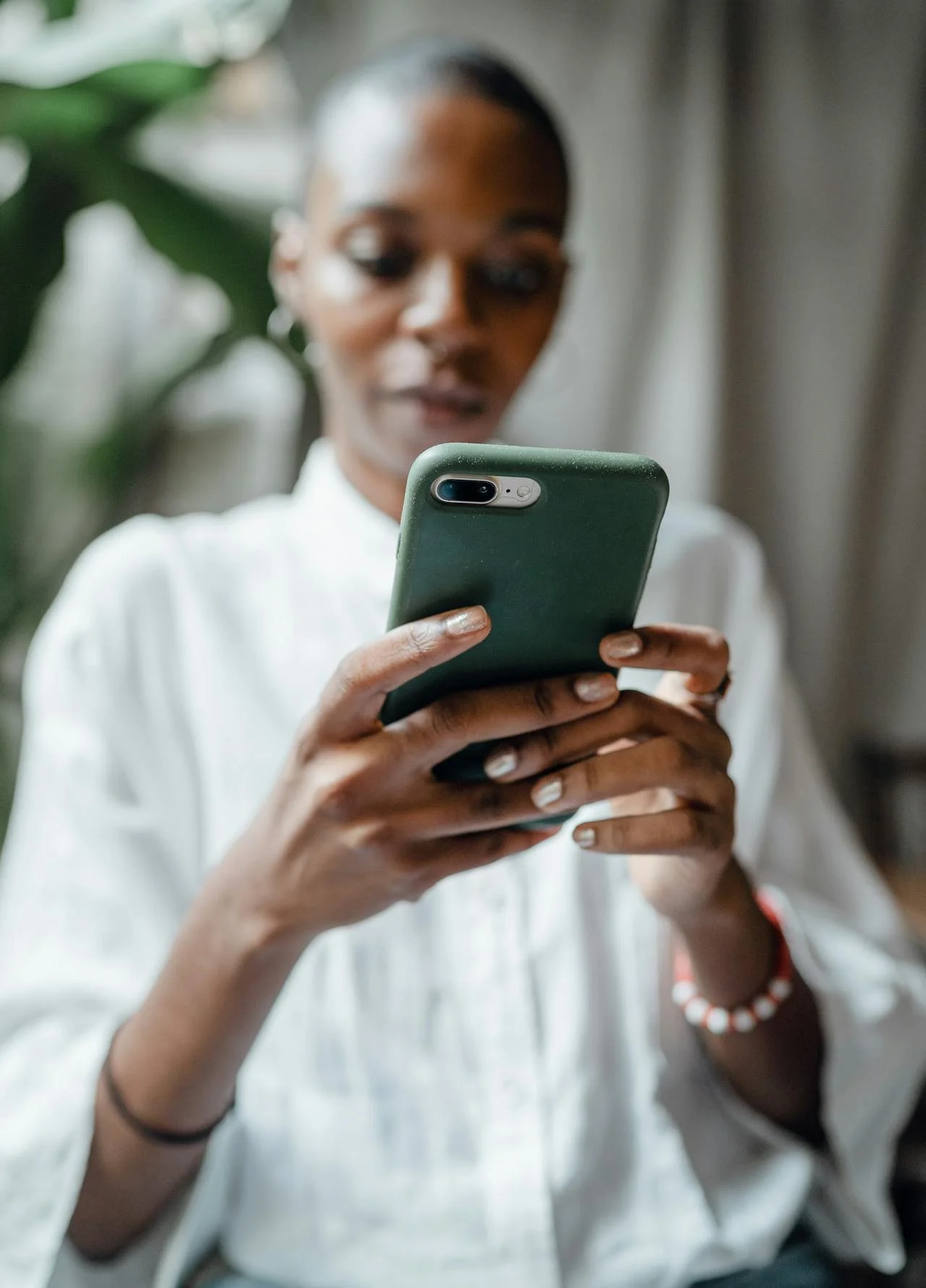 A woman with short hair and hoop earrings looking at her phone, wearing a white blouse, in front of a plant.