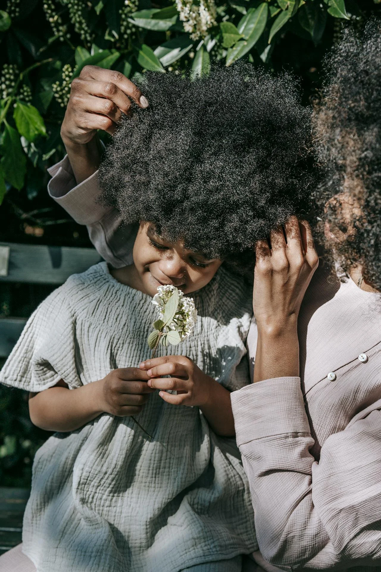 A woman and a young girl with curly hair are touching the girl's head, with the girl holding a white flower close to her face, in an outdoor setting with green foliage in the background.